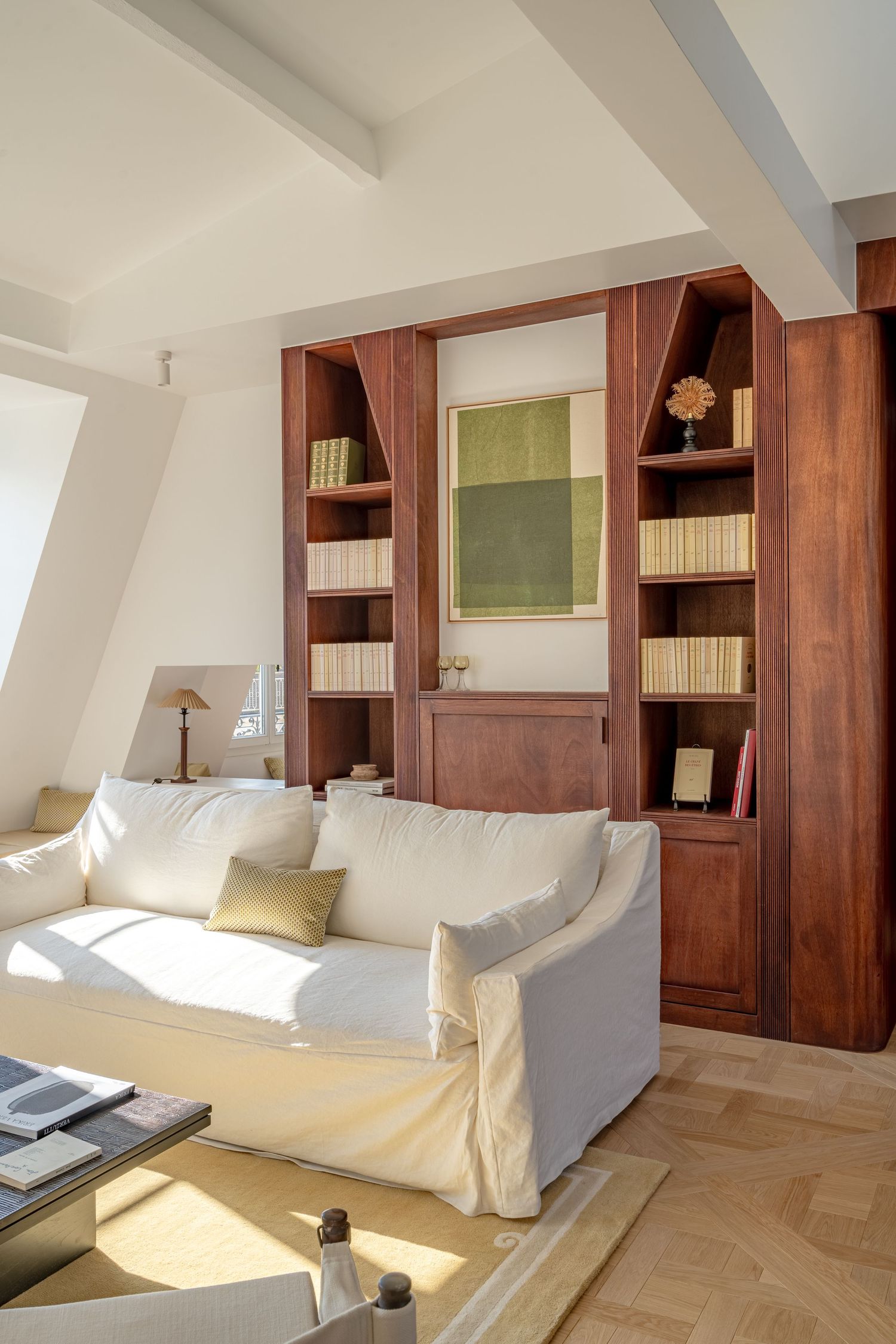 Bright living room with white sofa, mahogany built-in bookshelves, and sunlit herringbone wood floor.