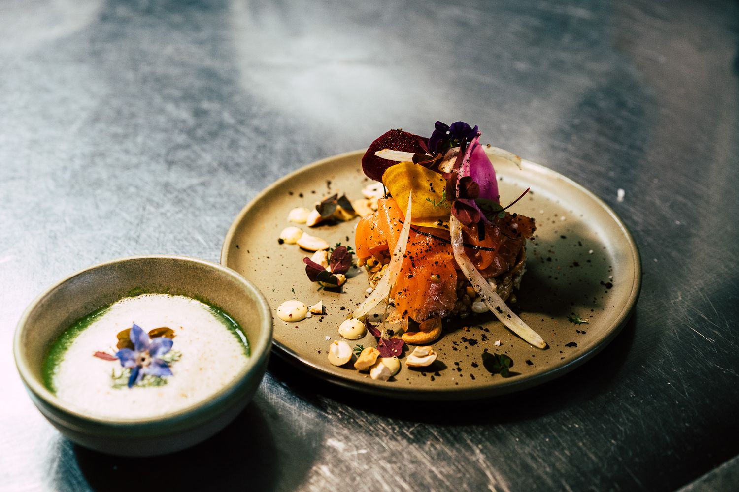 Fine dining gourmet dish with colorful garnishes served alongside a creamy soup on a metal surface.