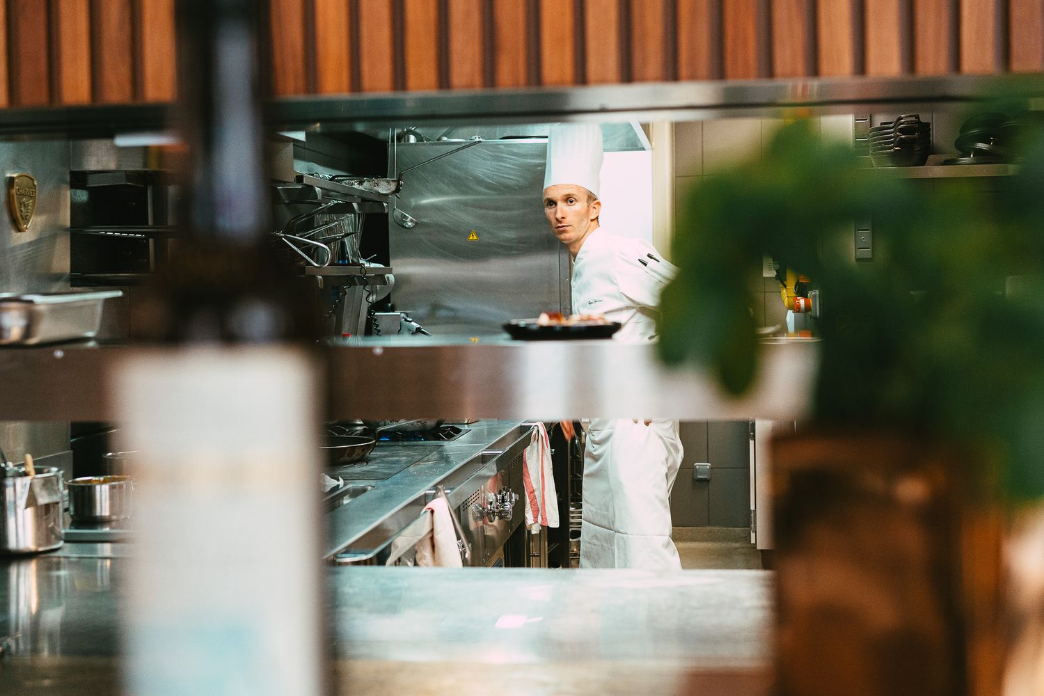 A chef in white uniform holding a pan in a professional restaurant kitchen viewed through a window.