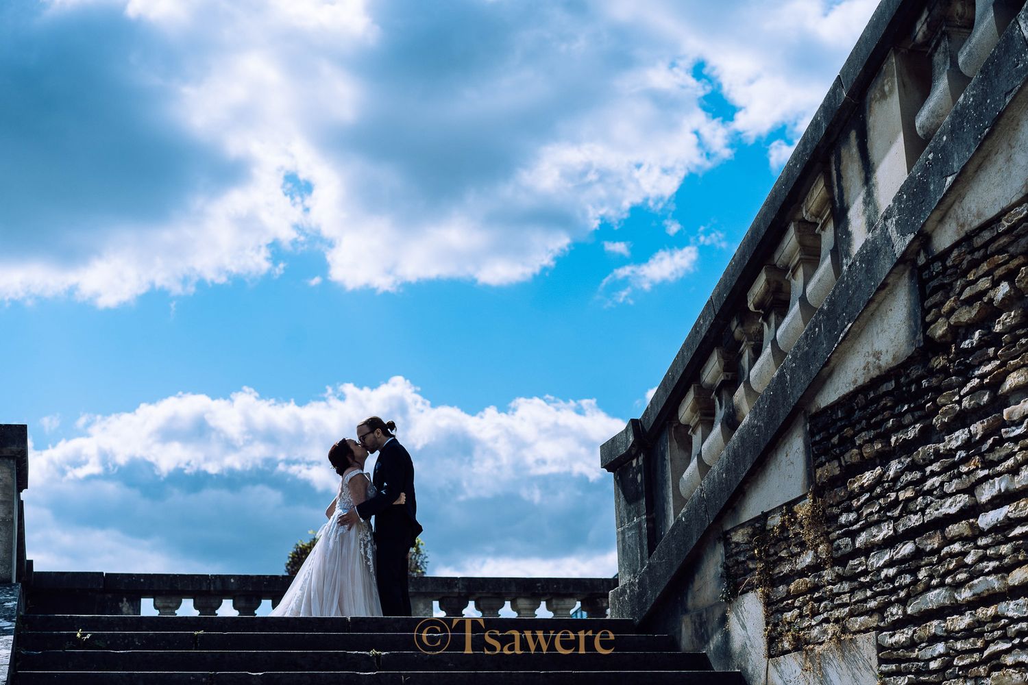 Bride and groom sharing a kiss on stone steps against a bright blue cloudy sky.