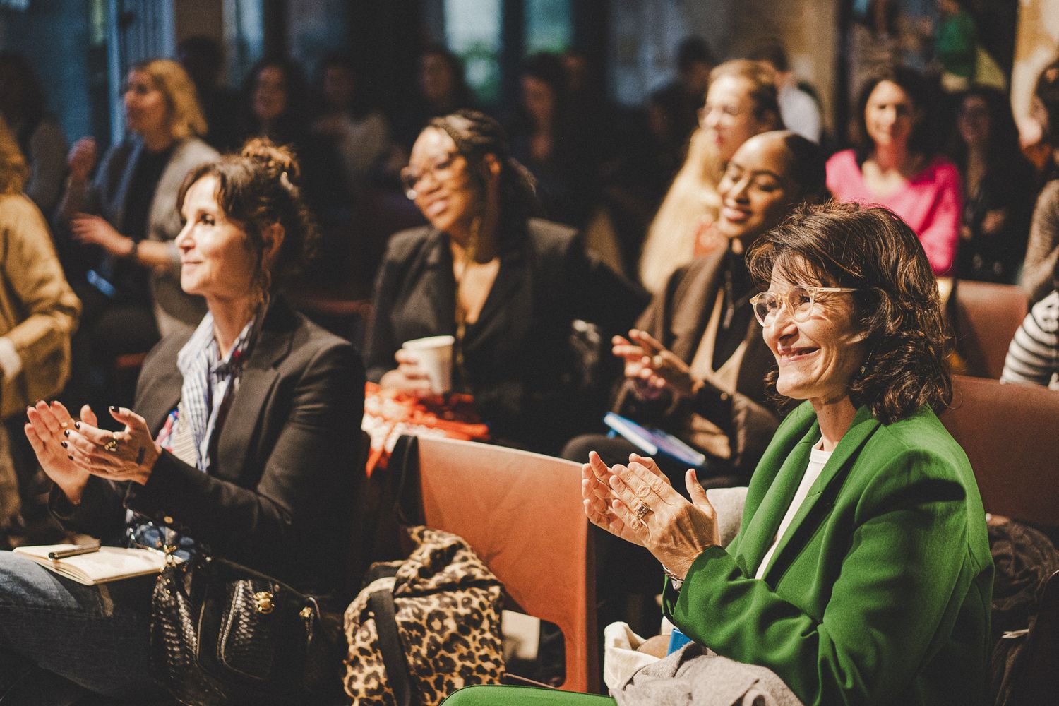 Women applauding at an indoor conference, one wearing a green blazer, seated in a warmly lit venue.