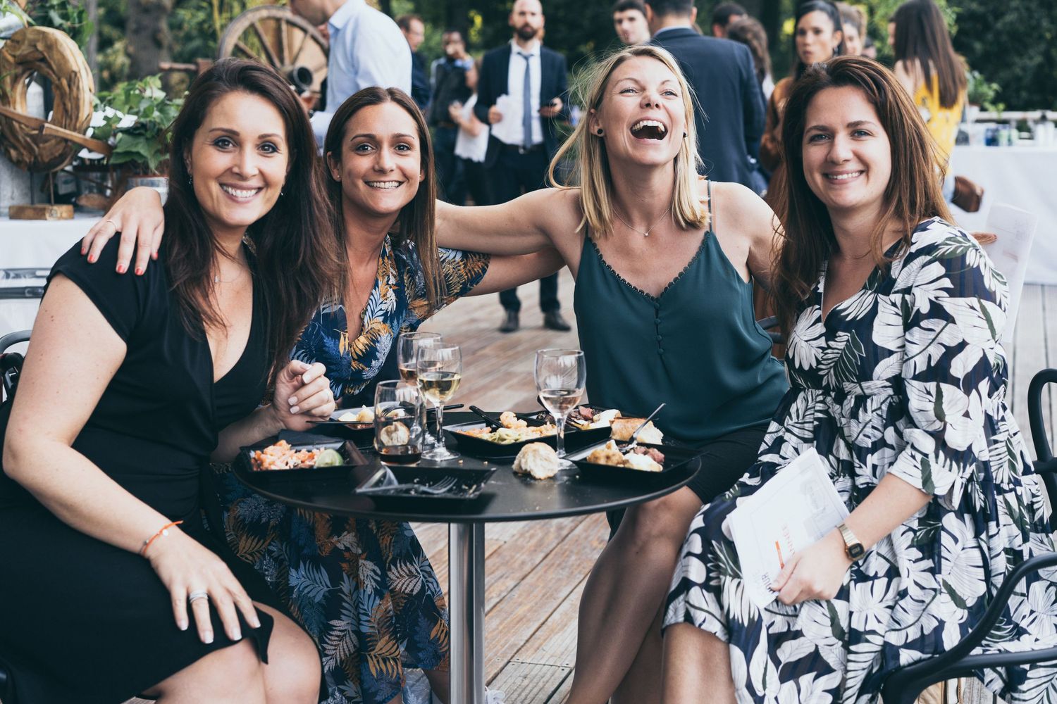 Four women smiling and posing together at an outdoor evening social gathering with drinks.