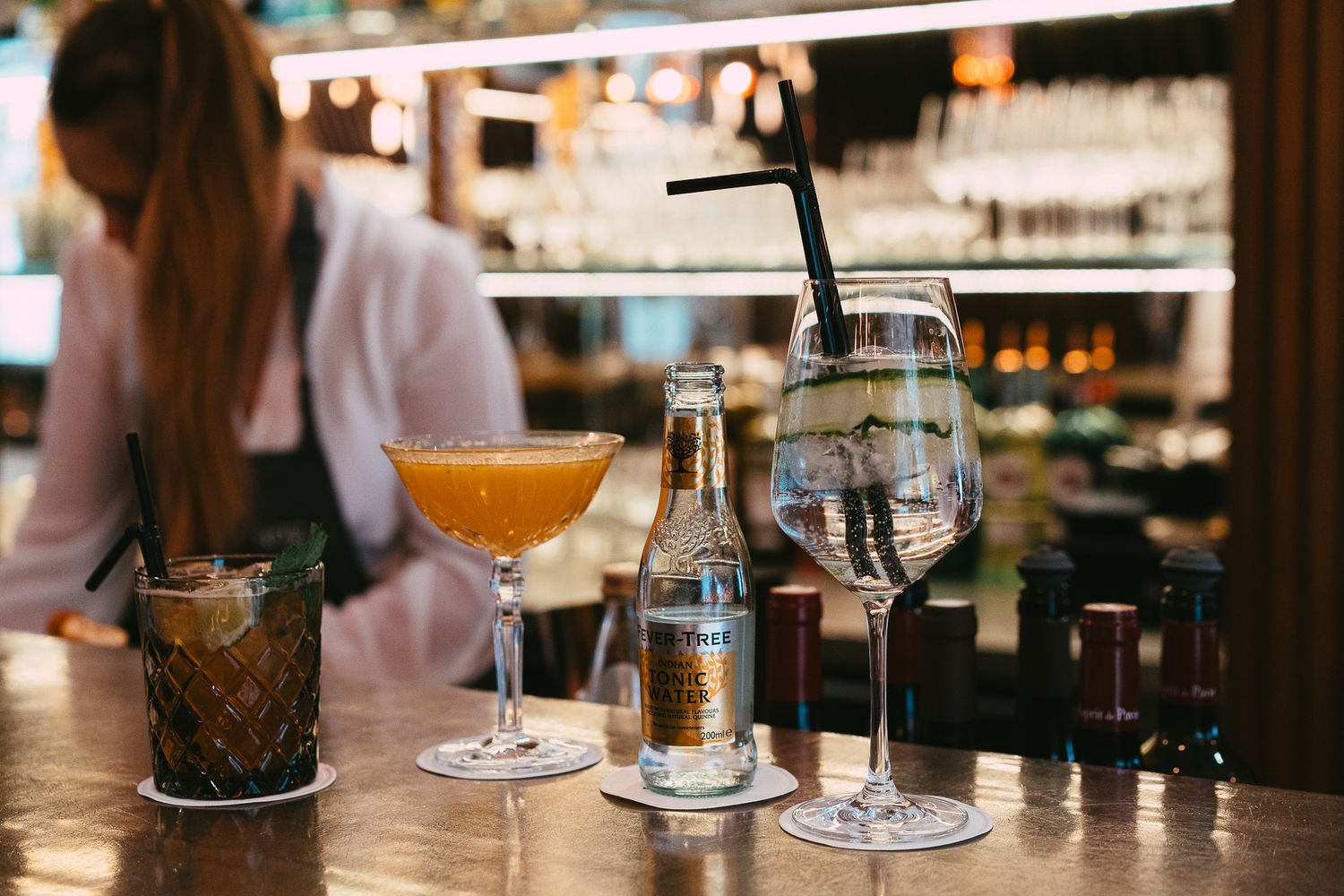 Bartender behind a bar counter with cocktails, a Fever-Tree bottle, and glassware in a dimly lit bar.
