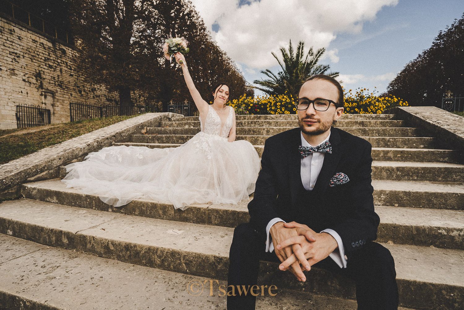Bride in ballgown celebrating on stone steps while groom sits in foreground at outdoor wedding.