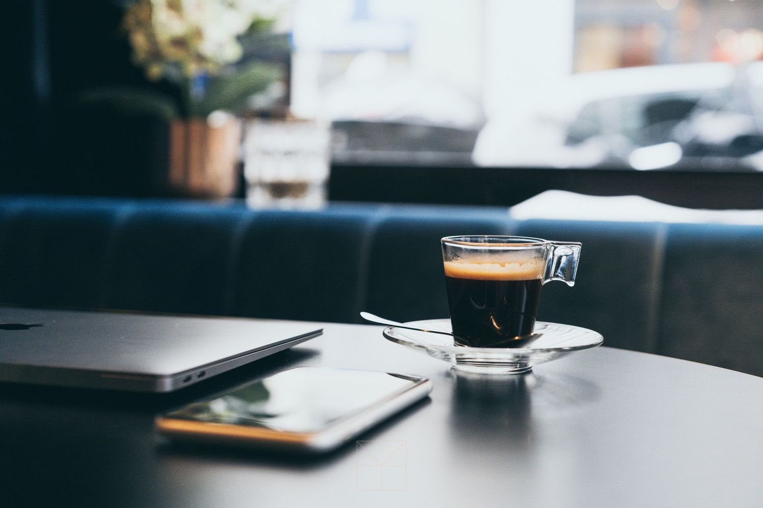Glass espresso cup on a saucer next to a laptop in a modern cafe setting.