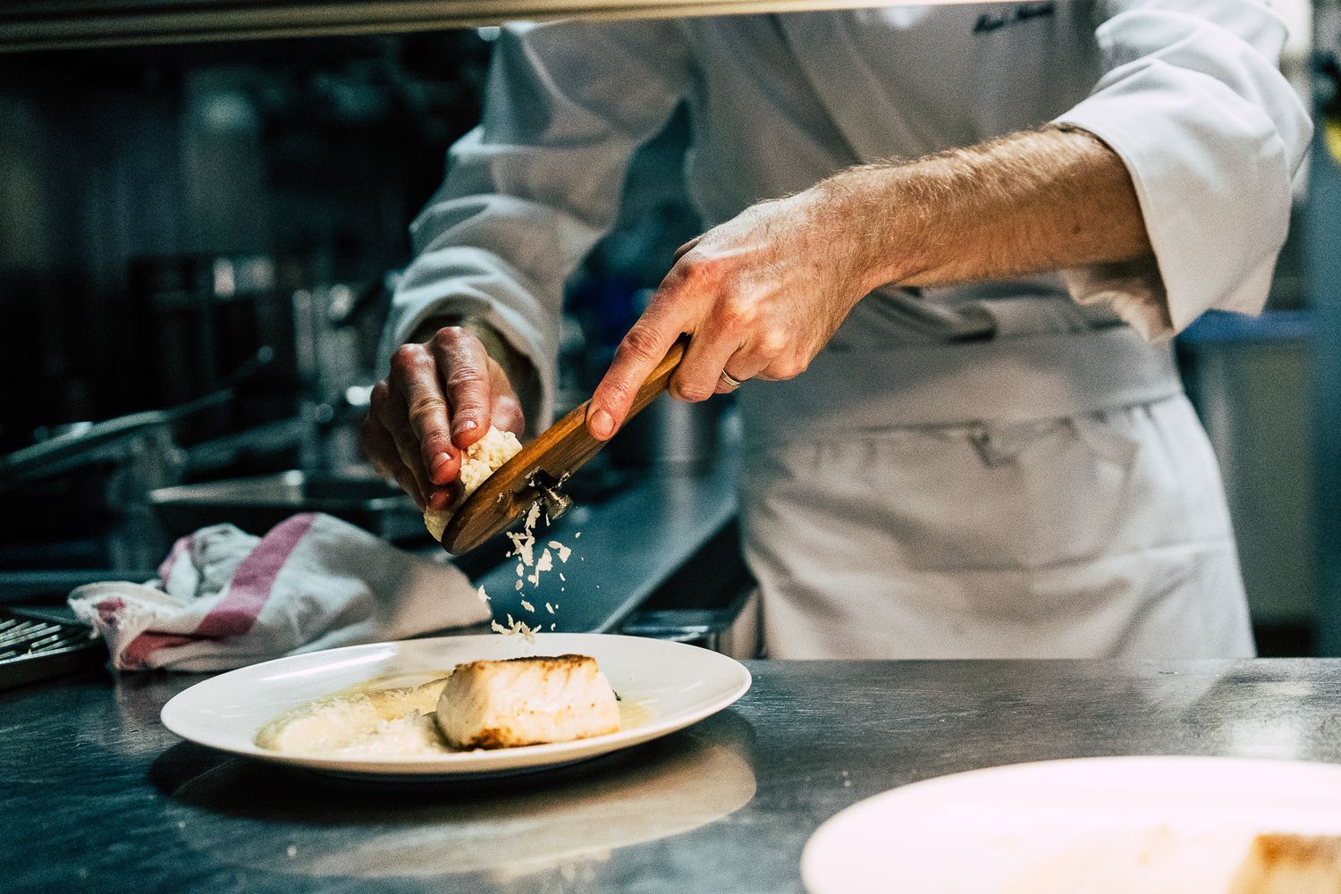 Chef seasoning a fish fillet on a white plate in a professional restaurant kitchen.