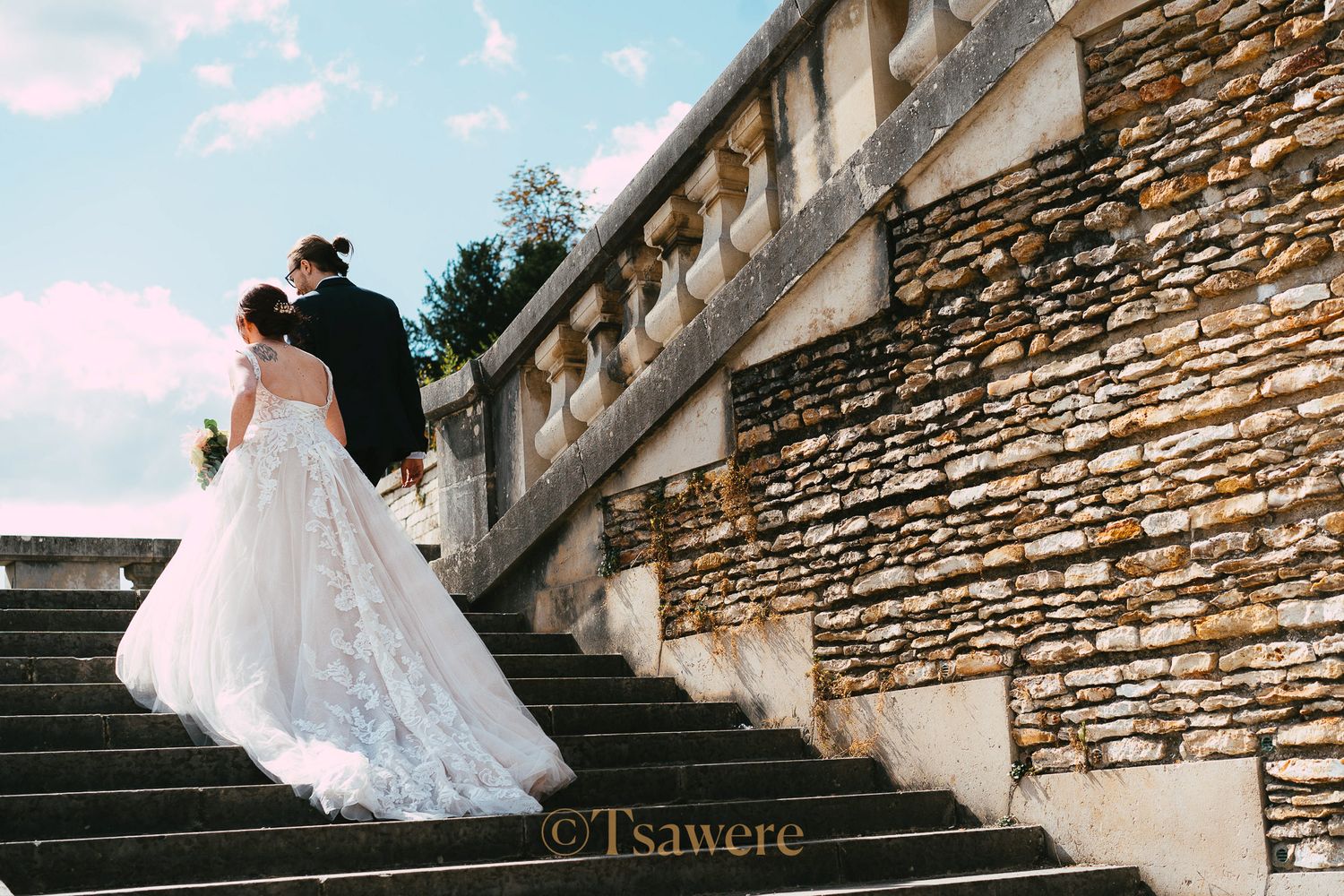 Bride and groom walking up stone steps at elegant outdoor wedding venue under blue sky.