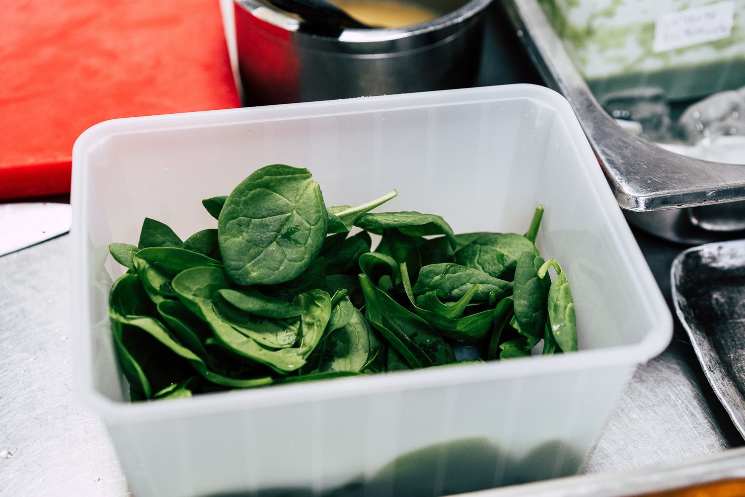 Fresh baby spinach leaves in a white plastic container on a stainless steel kitchen surface.
