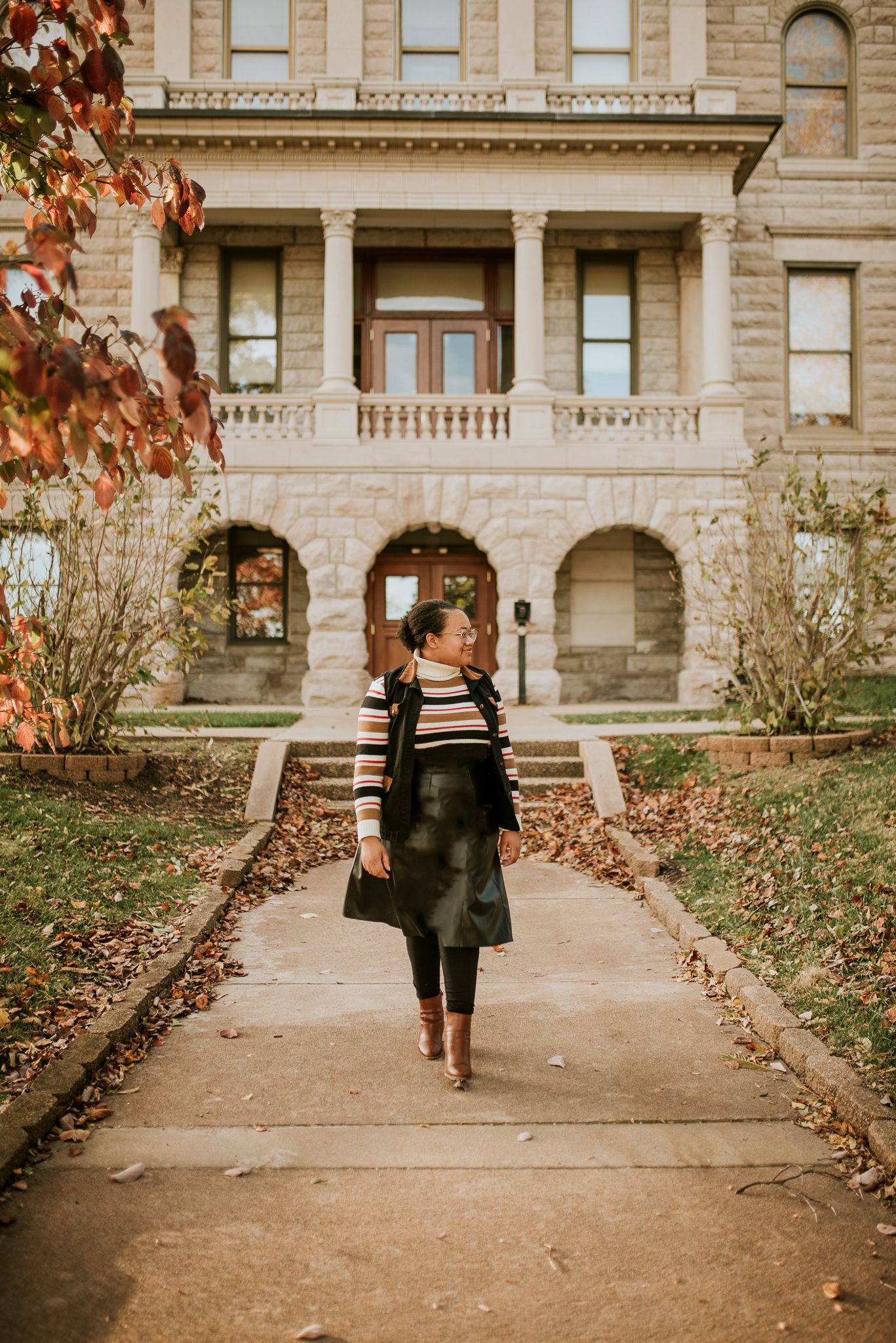 Woman in striped sweater and black skirt walking on a path in front of a historic stone building in autumn.