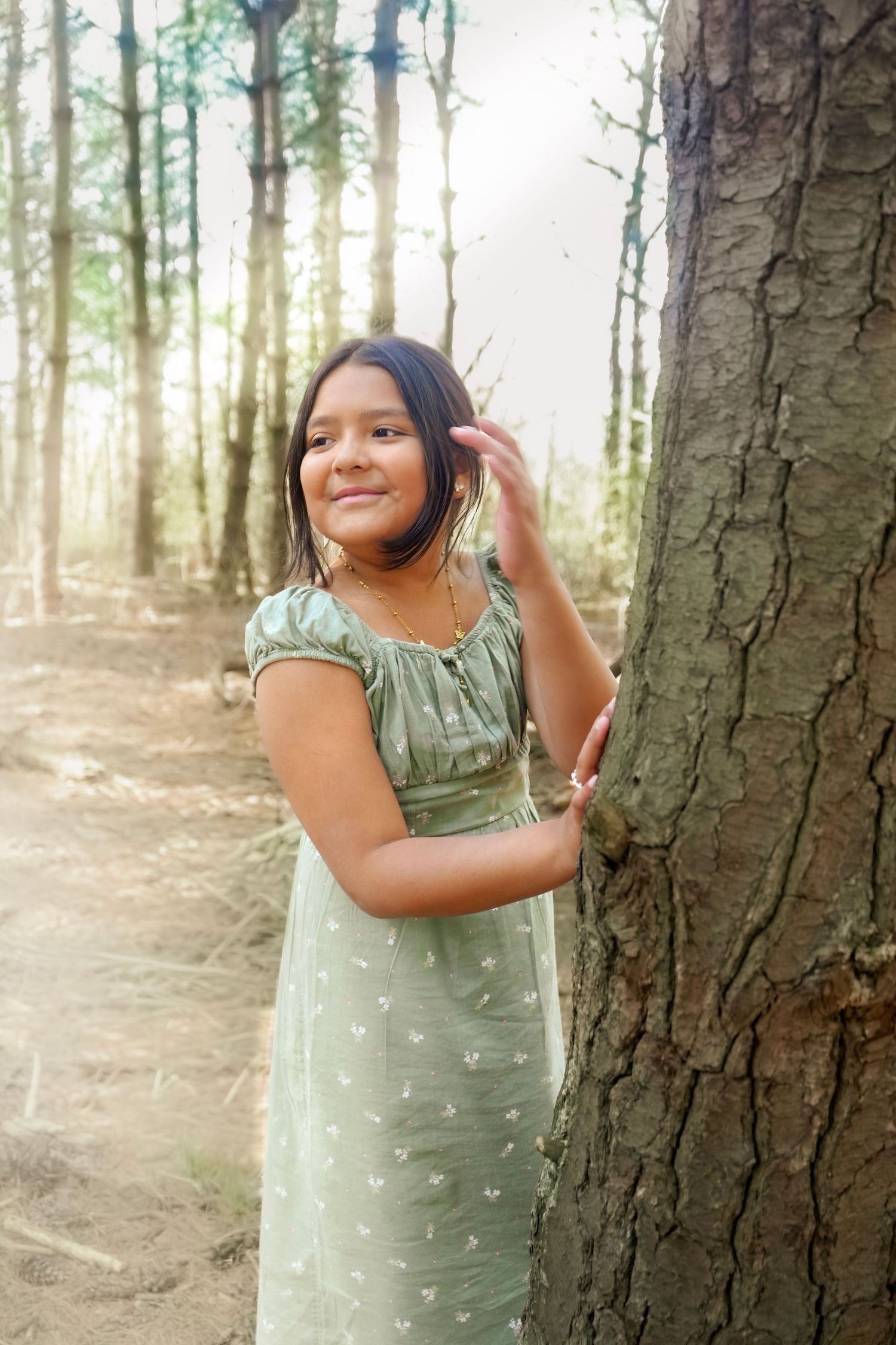 Young girl in green floral dress smiling beside a tree trunk in a sunlit forest.