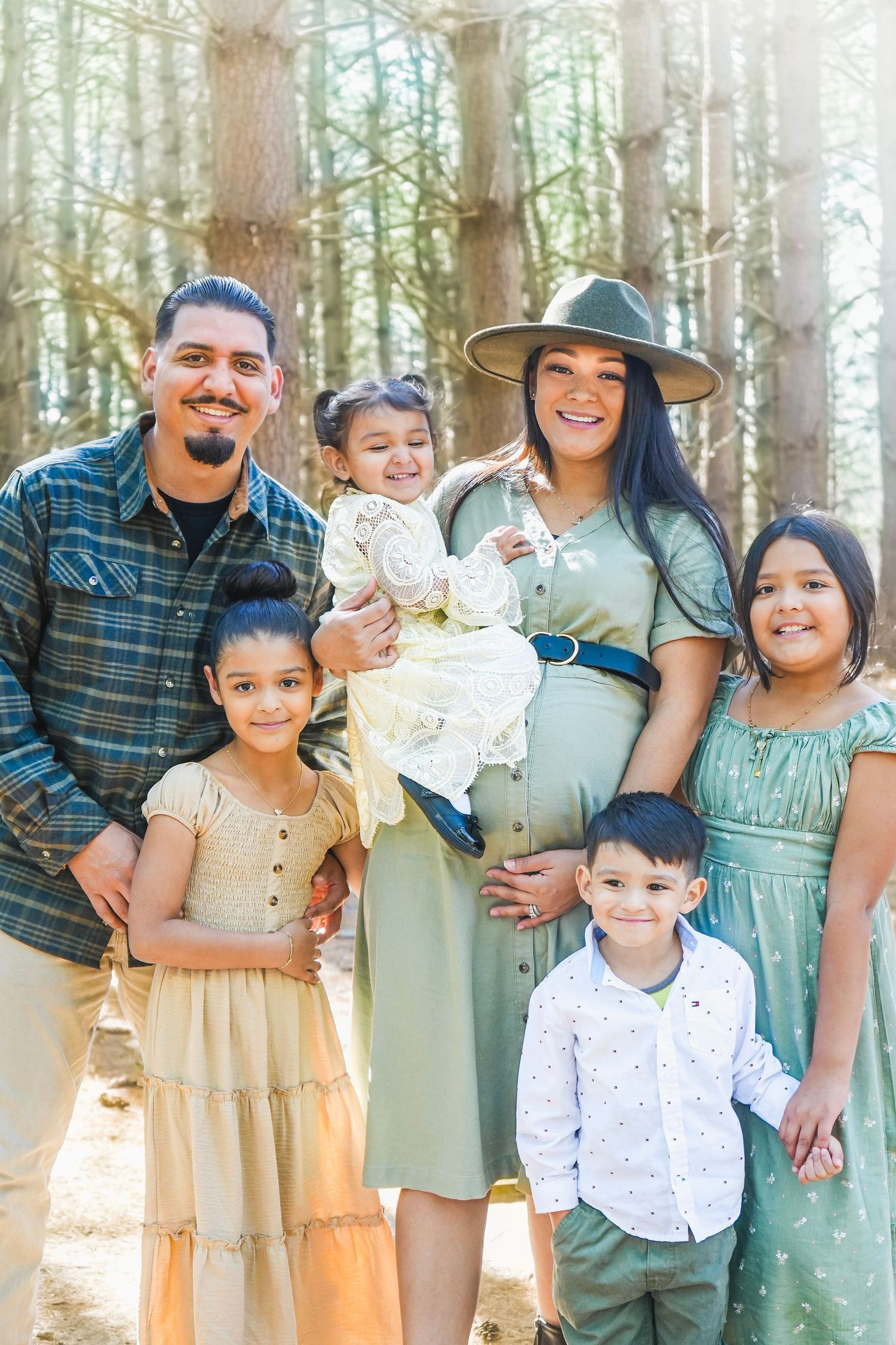 A happy family of six poses in a sunlit forest, with parents and four young children dressed in coordinating green and neutral outfits.
