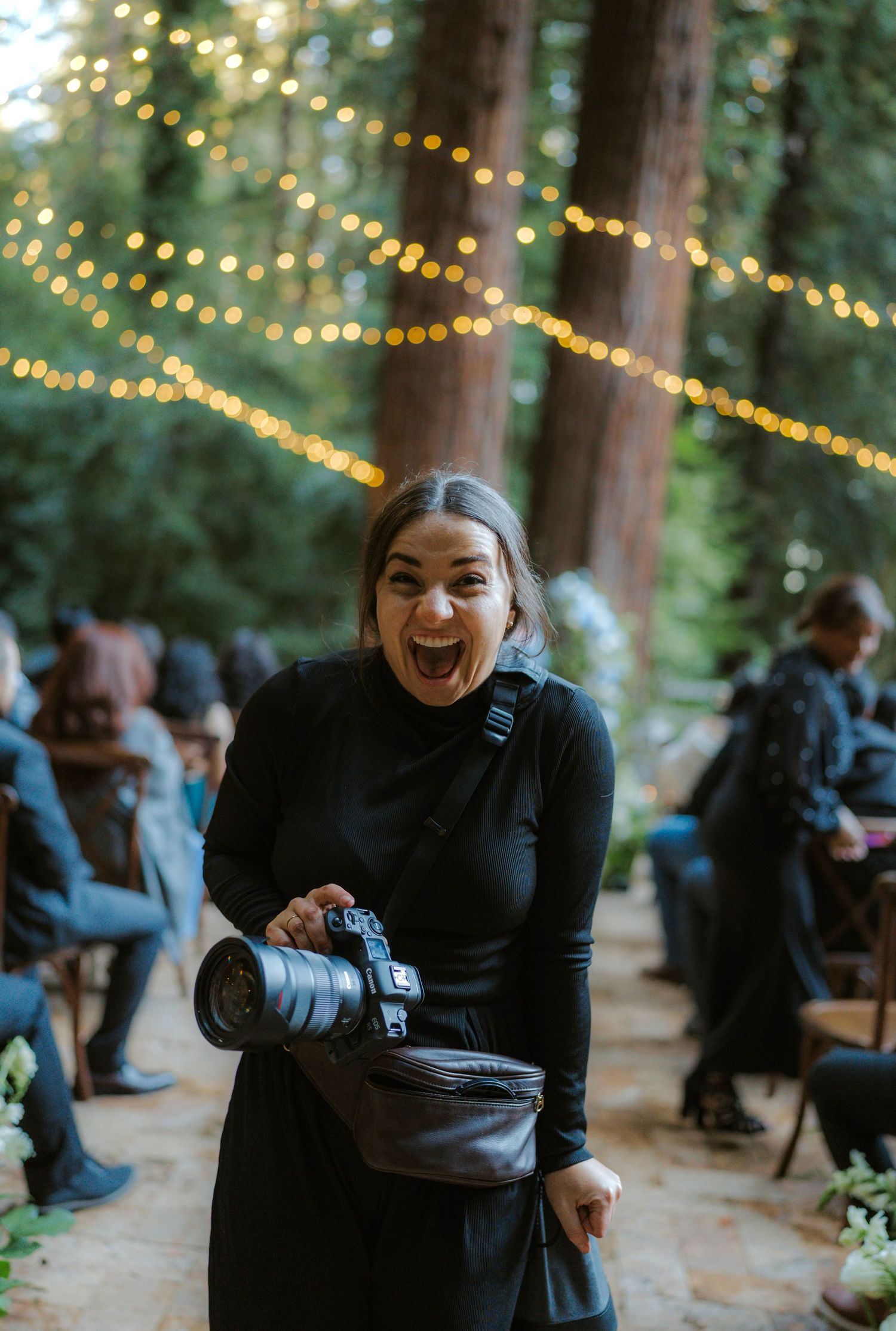 Person in black outfit laughing joyfully at outdoor event with twinkling string lights in forest setting.