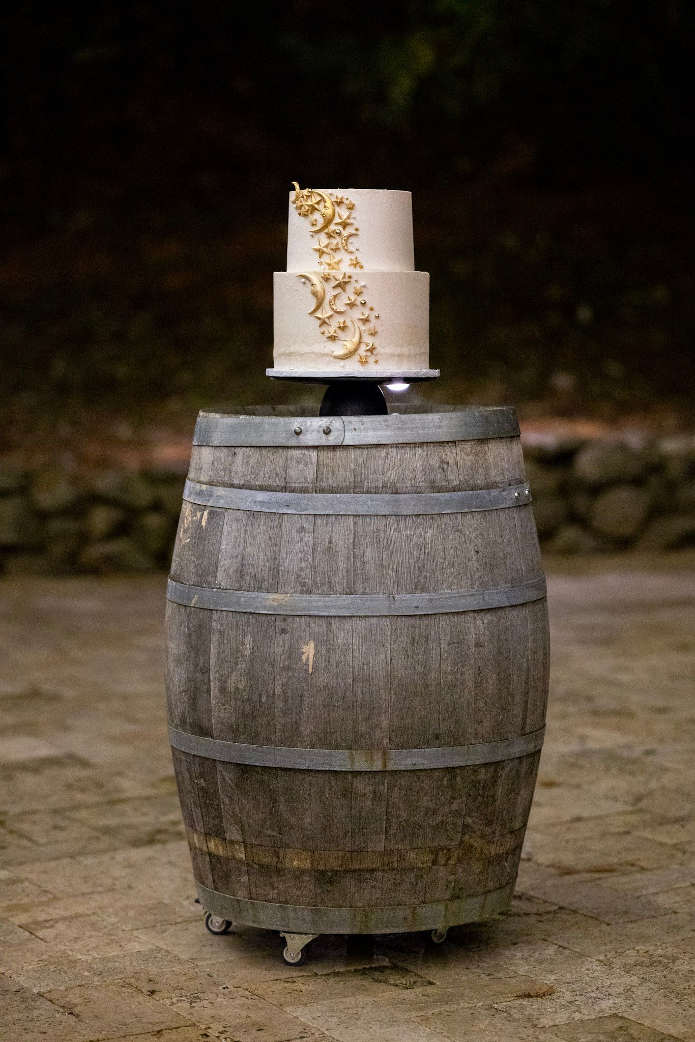 A simple two-tier white wedding cake with gold leaf details displayed on top of a rustic wooden wine barrel outdoors.