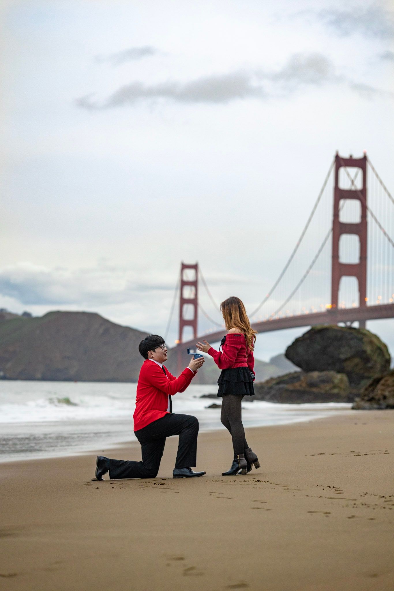 A romantic proposal scene at Baker Beach with the iconic Golden Gate Bridge in the background on a cloudy day.