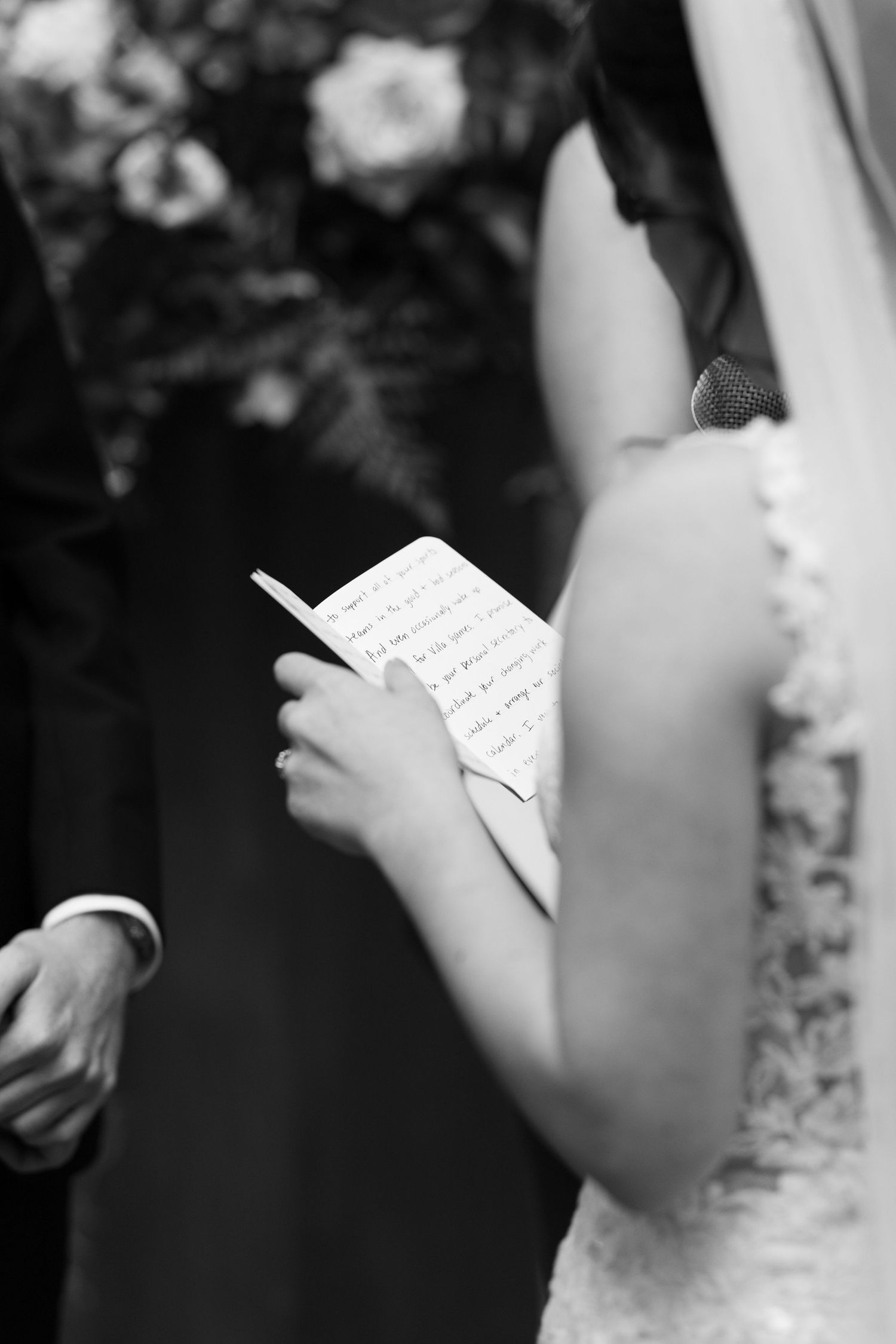 Bride reading handwritten wedding vows from a card during ceremony in elegant lace gown, black and white photo.