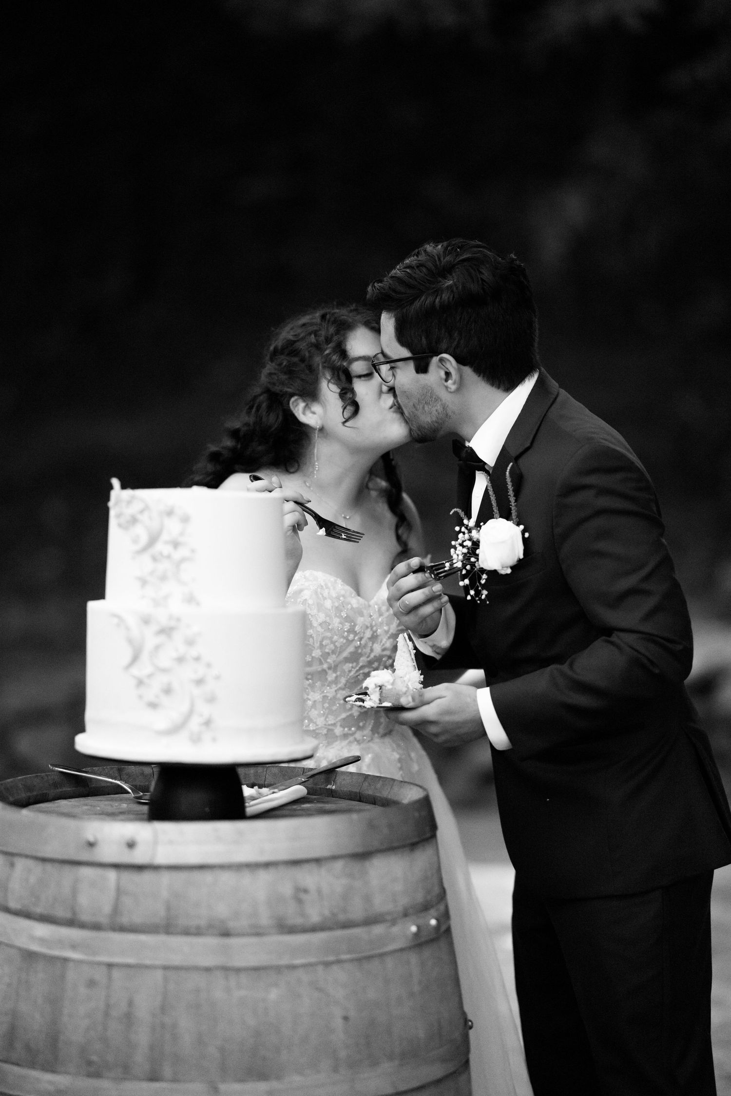 Black and white wedding photo series showing couple kissing behind wedding cake placed on rustic wine barrel.