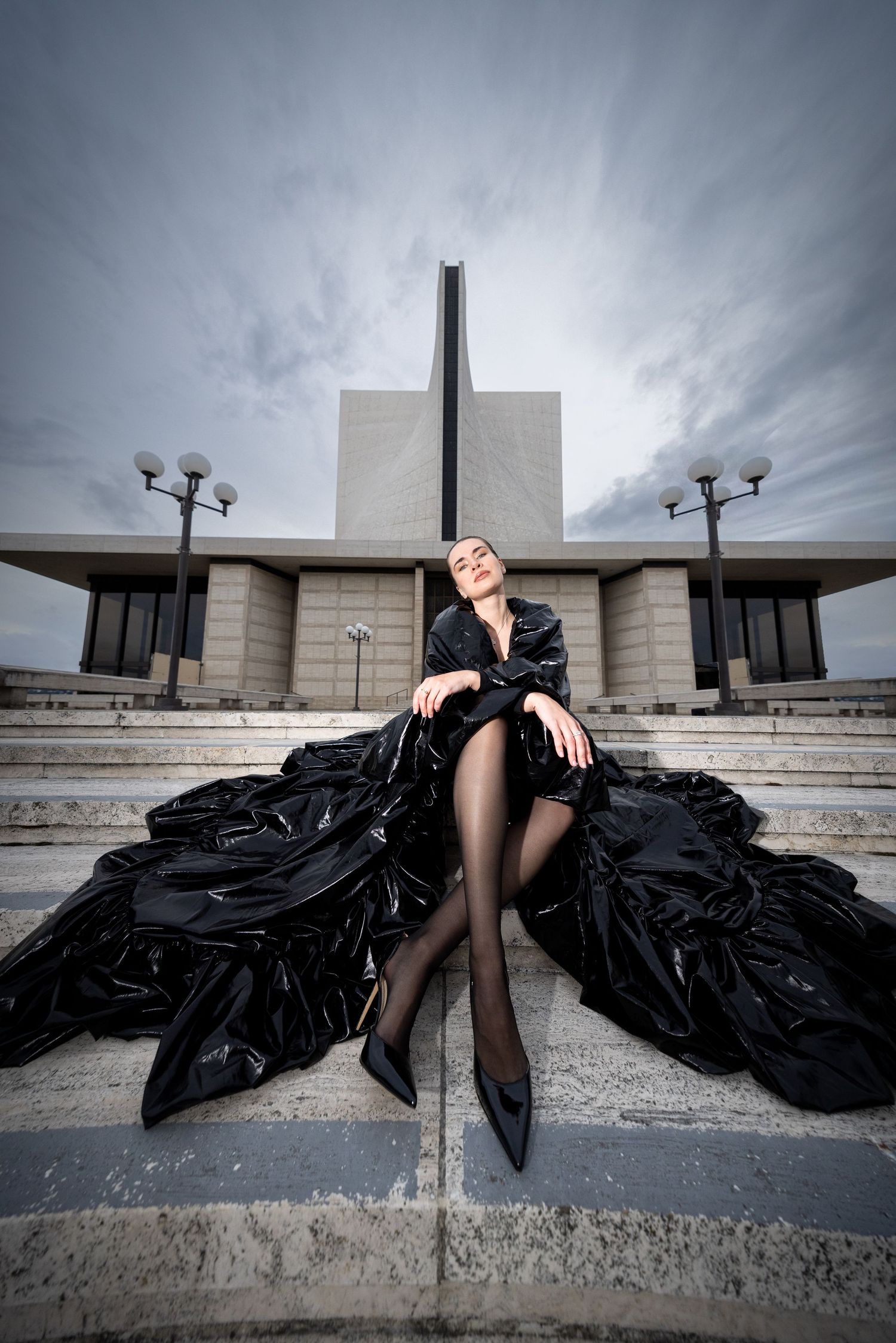 Woman in dramatic black gown sitting on steps before a modernist monument under a cloudy sky.