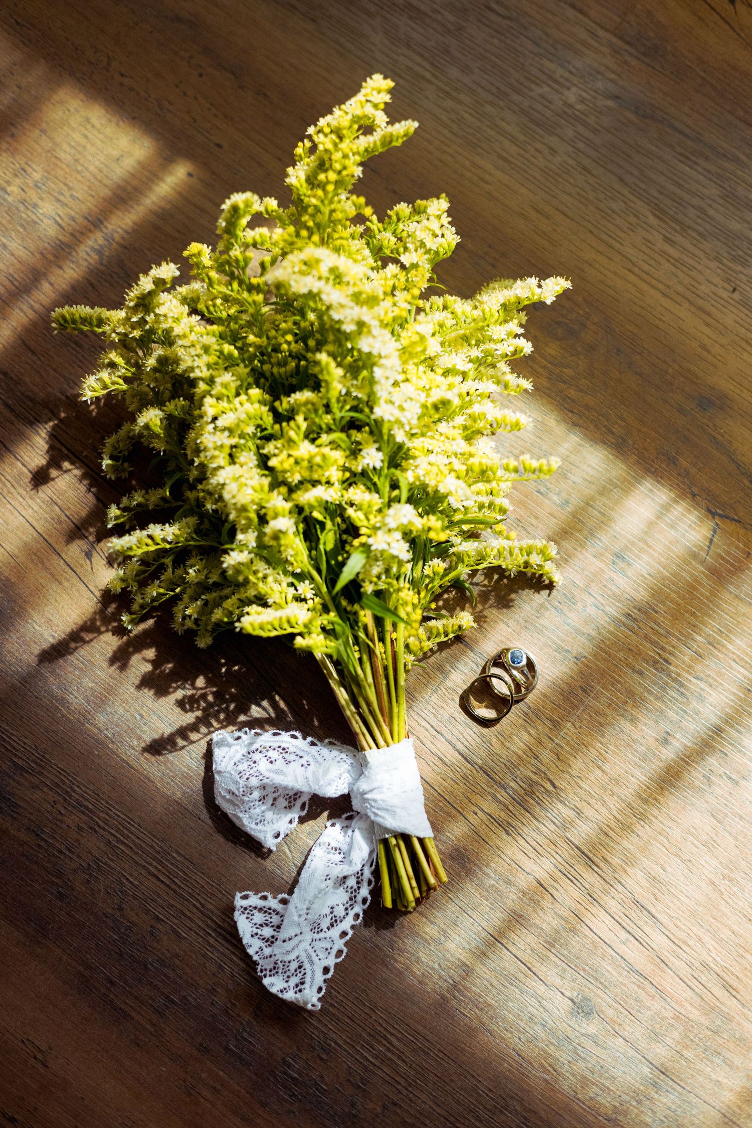 Yellow wildflower bouquet tied with white lace ribbon beside wedding rings on rustic wooden floor in sunlight.