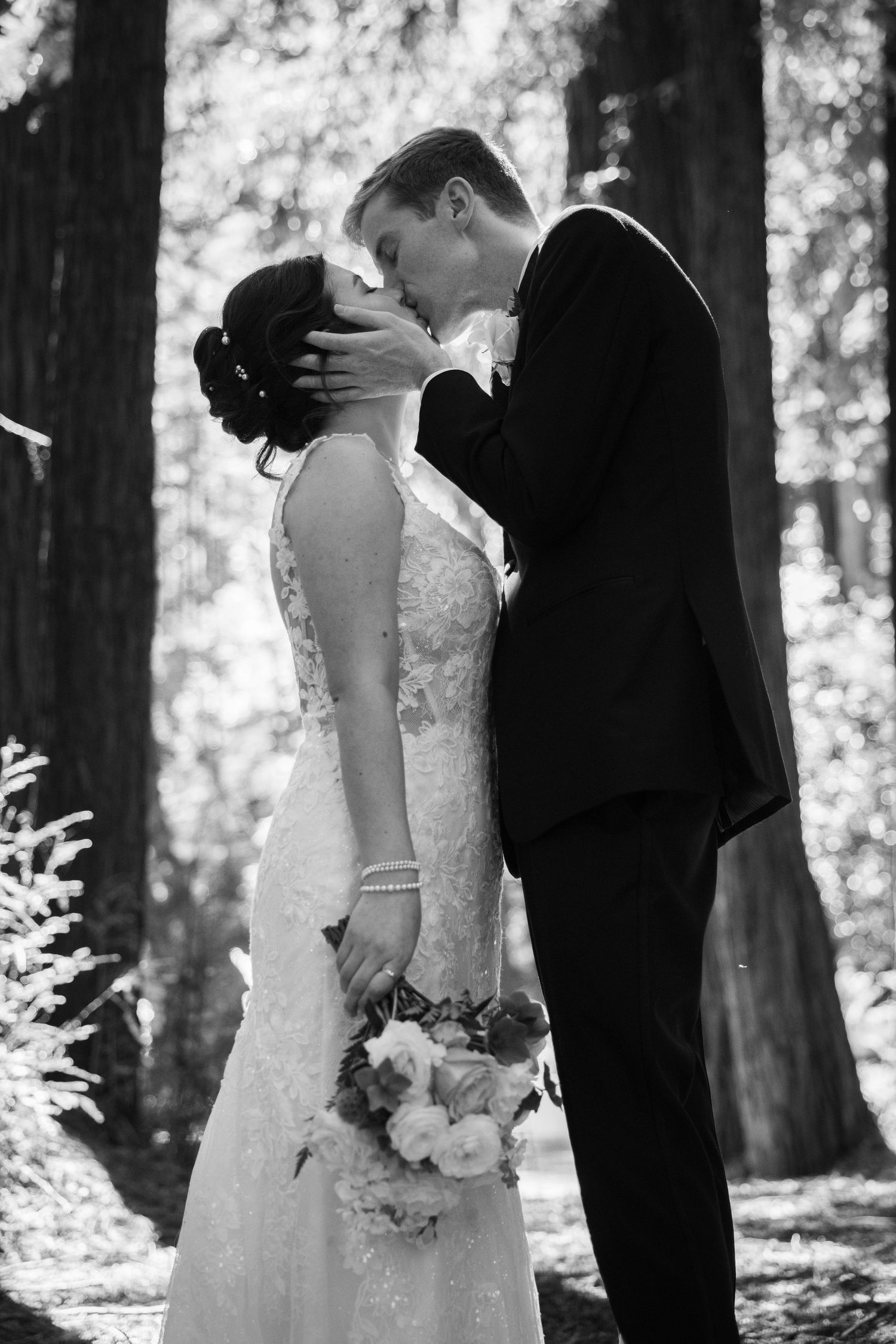 Black and white photo of a bride and groom sharing a romantic kiss in a forest, bride holding a bouquet.