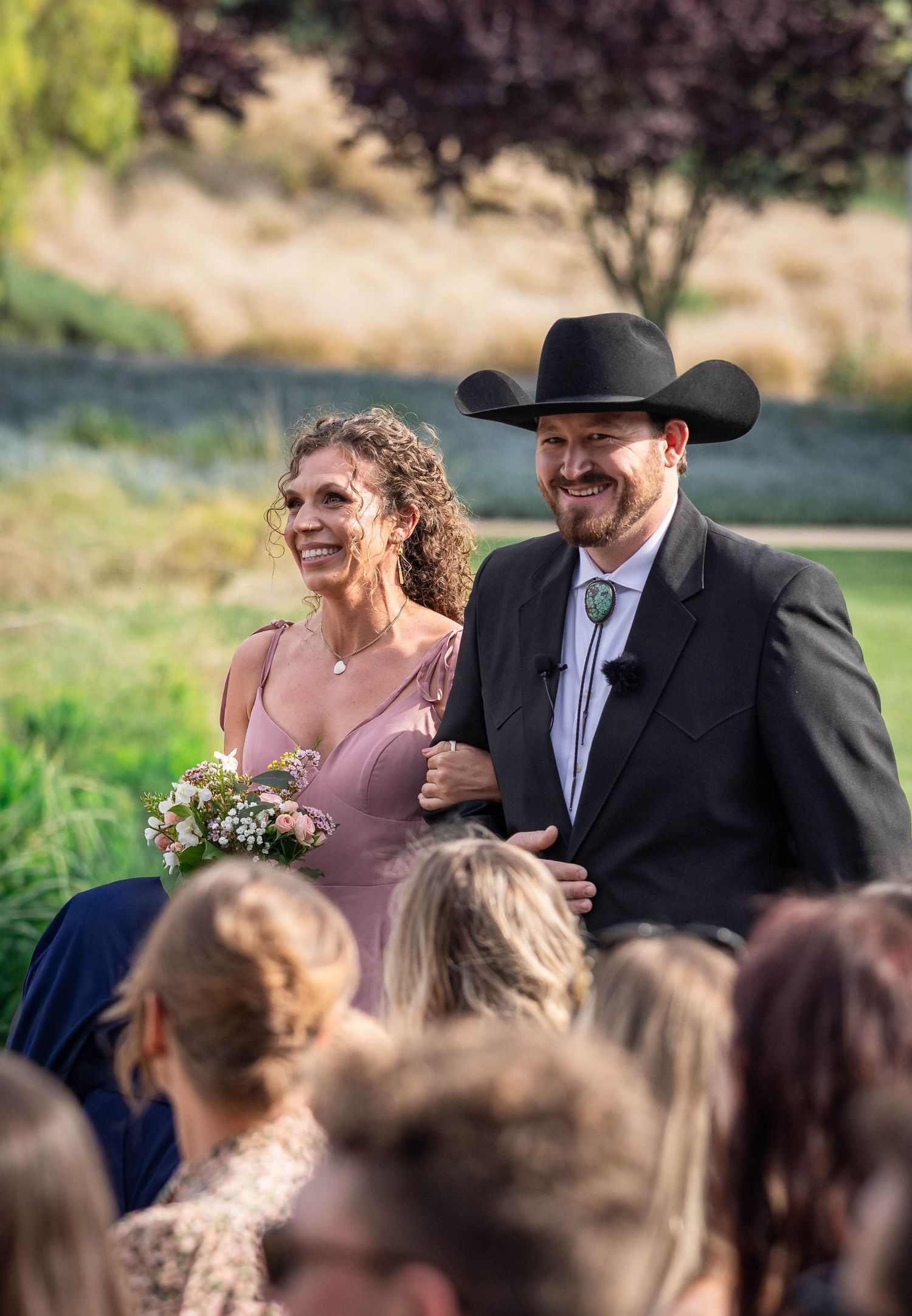 A smiling couple walks down the aisle at an outdoor wedding, man wearing a black cowboy hat and suit.