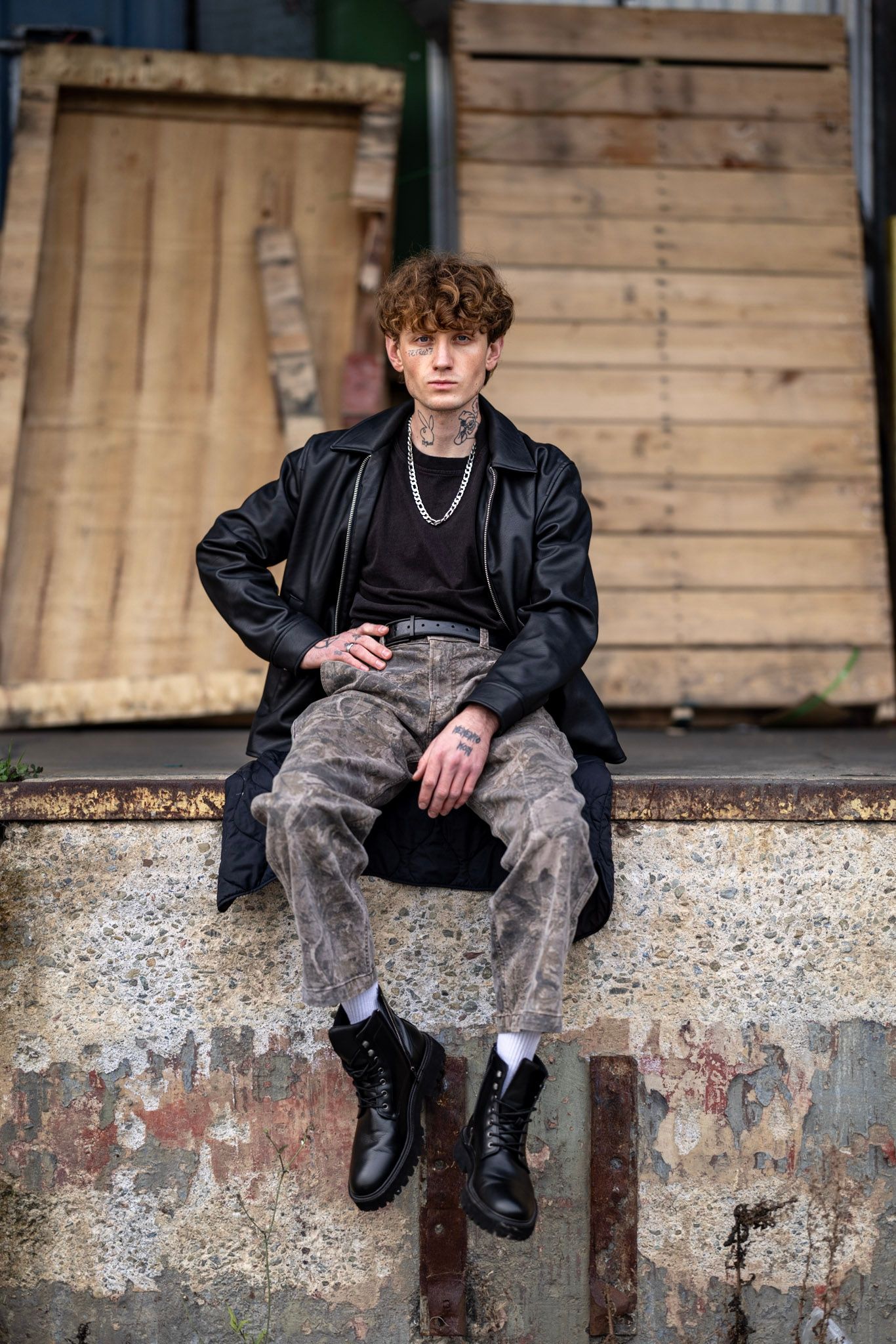 A person in a black jacket and distressed pants sits on a concrete ledge against a weathered wooden wall.