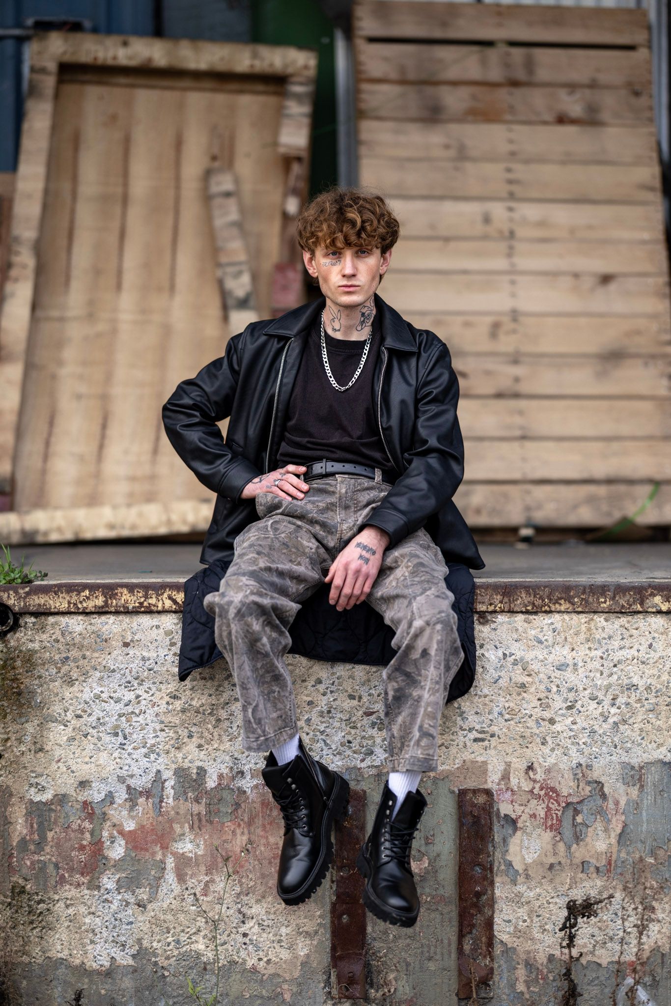 A person in a black leather jacket and camo pants sits on a concrete ledge wearing combat boots and a silver necklace.