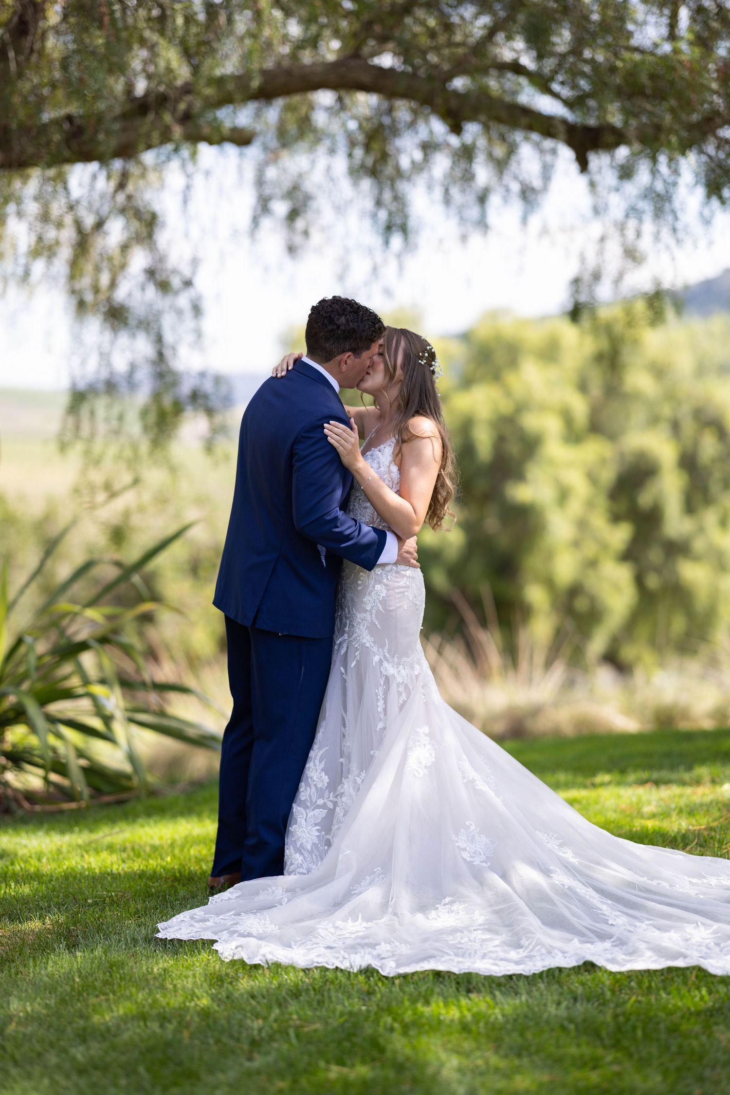 A romantic wedding couple shares a tender moment on lush grass under drooping willow trees on their special day.