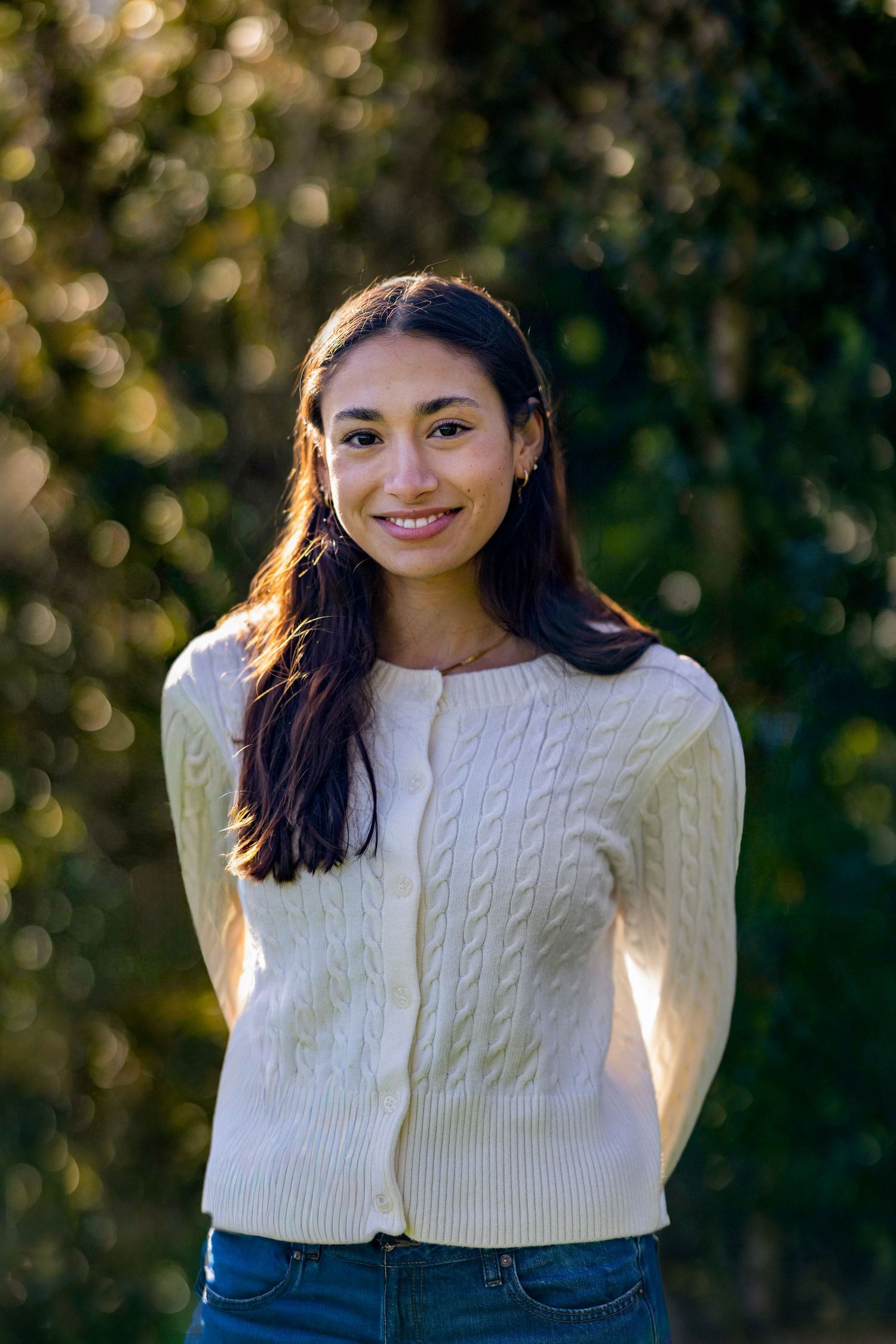 A person in a white cable knit sweater and jeans poses outdoors with a natural bokeh background.