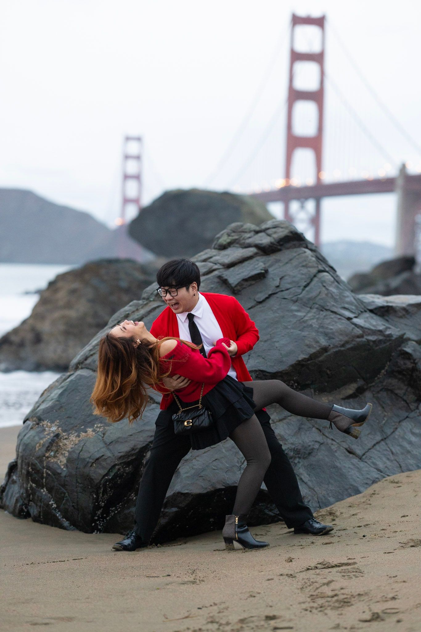 A couple in red and black outfits dance passionately on a beach near the Golden Gate Bridge in San Francisco.