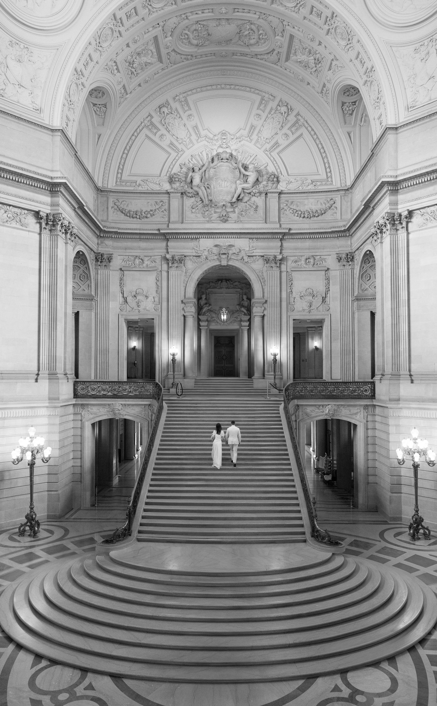 Black and white photo of a couple ascending grand staircase in ornate Beaux-Arts building interior.