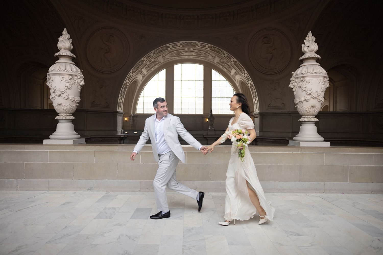 Newlywed couple holding hands and walking through an elegant domed hall, bride in white gown with bouquet.