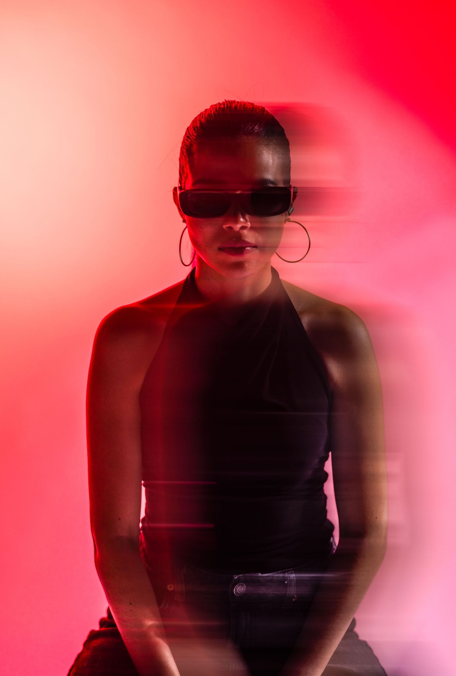 Woman in black halter top and oversized sunglasses poses against vibrant red background with dramatic motion blur effect.