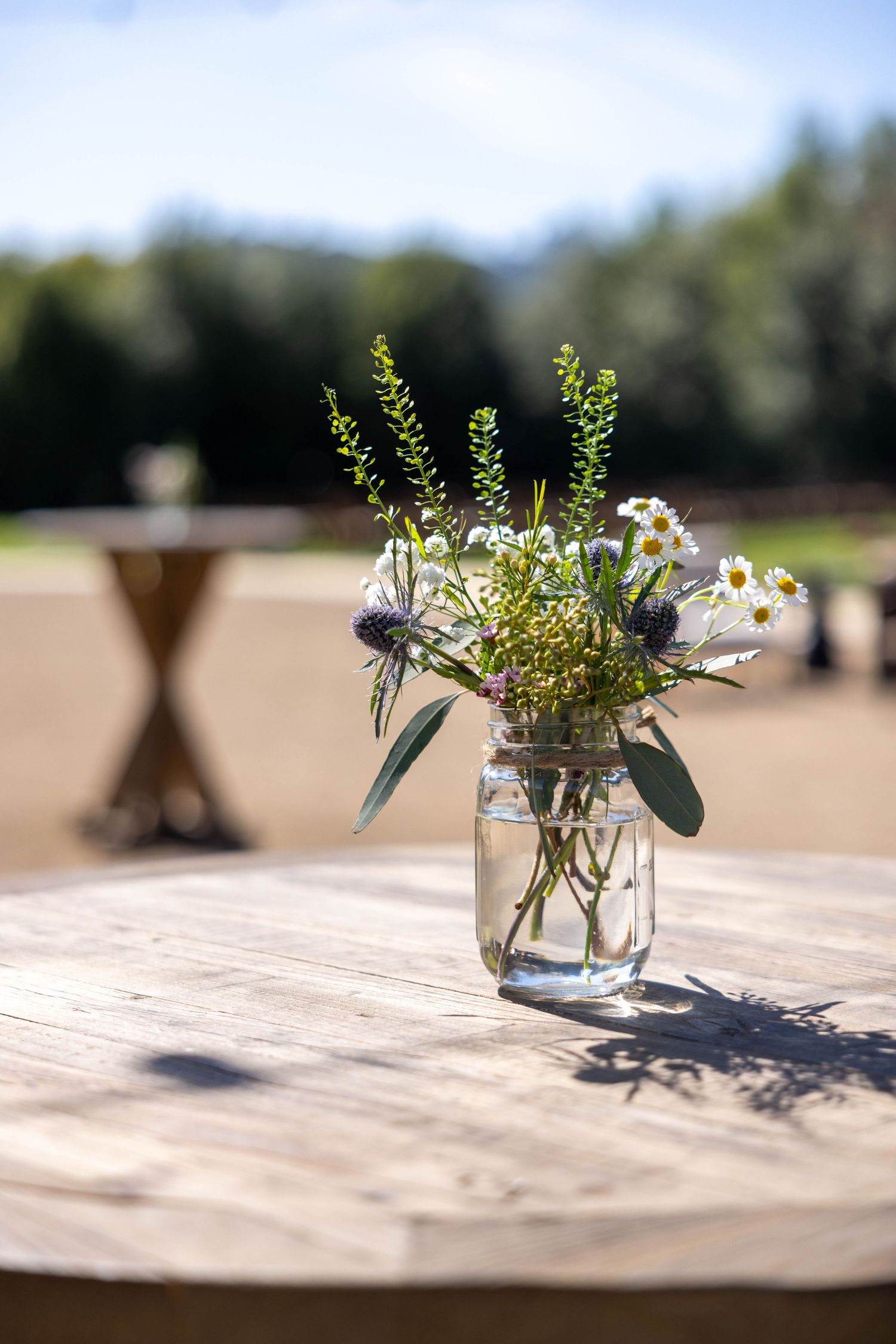 A rustic mason jar centerpiece with delicate wildflowers and greenery sits on a wooden table in natural outdoor lighting.