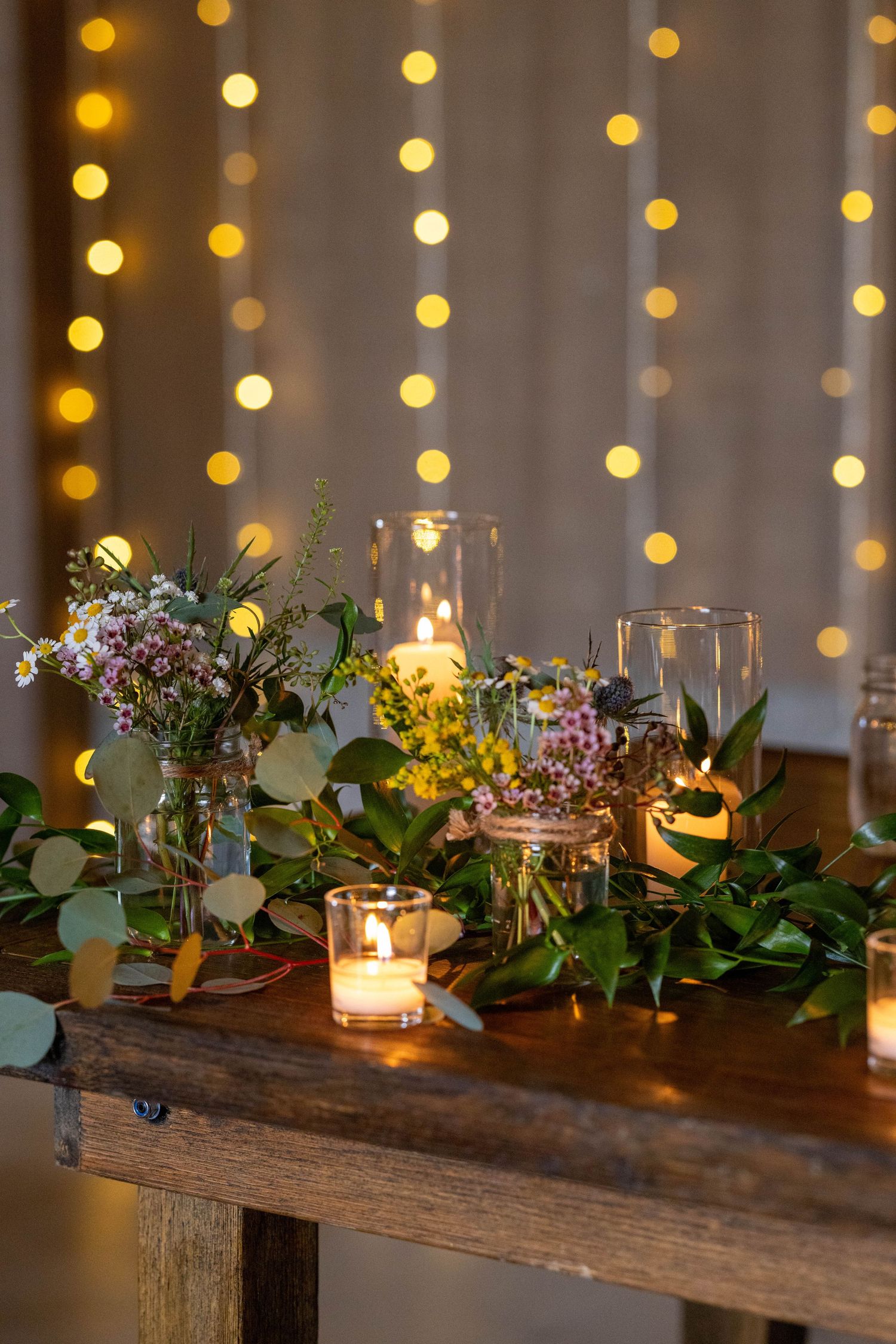 Rustic wooden table centerpiece with glowing candles and greenery against a backdrop of twinkling string lights.