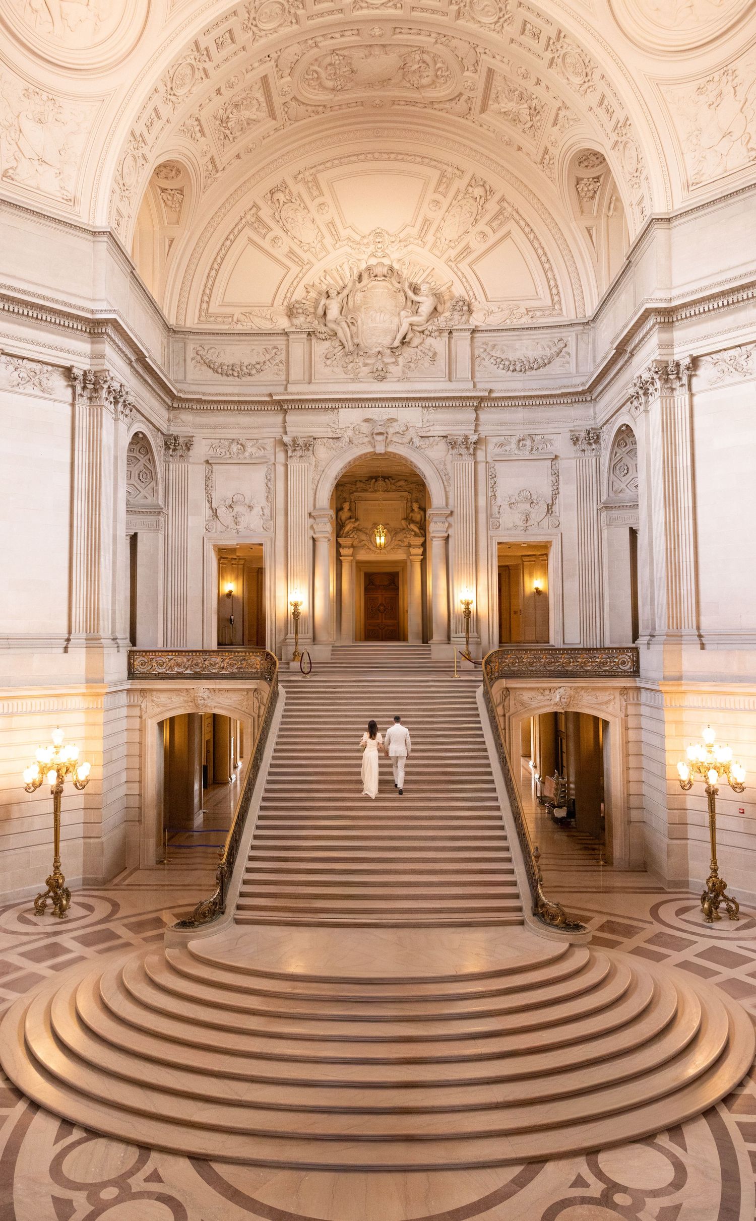 Couple ascending grand staircase in San Francisco City Hall's ornate Beaux-Arts rotunda with elegant marble architecture.