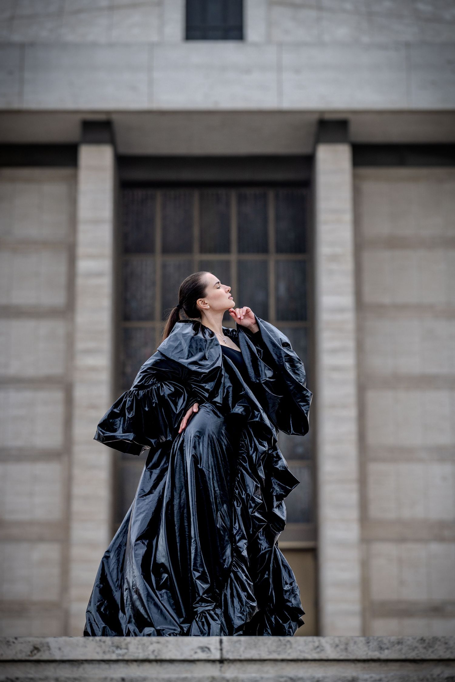 Woman in dramatic black glossy gown poses on stone steps of neoclassical building.