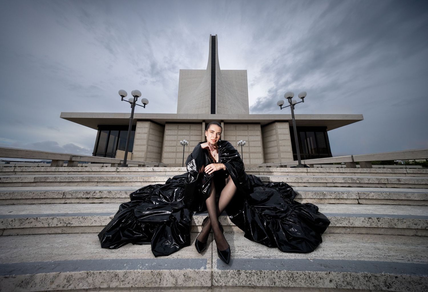 Woman in dramatic black gown poses on grand stone steps before a monumental brutalist building under cloudy skies.