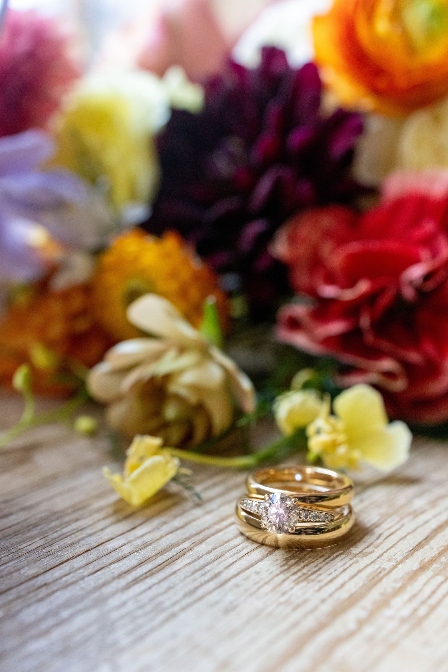 Gold wedding rings with diamond accents rest on a wooden surface before a vibrant colorful floral bouquet.