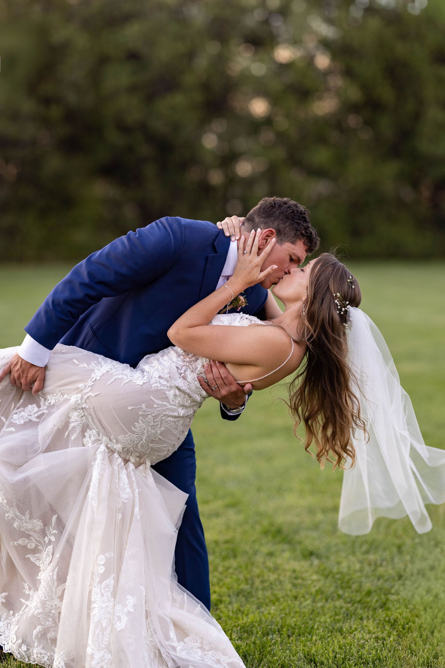 A couple in wedding attire performs a romantic dip pose on a grassy lawn during their outdoor photo session.