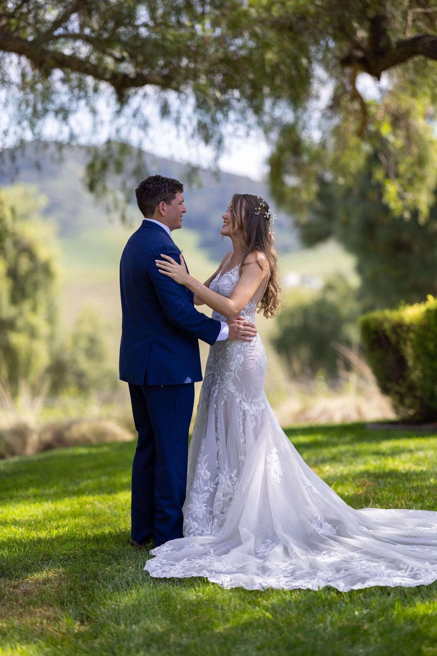 Newlywed couple sits together on grassy hillside under trees during romantic outdoor wedding photoshoot.