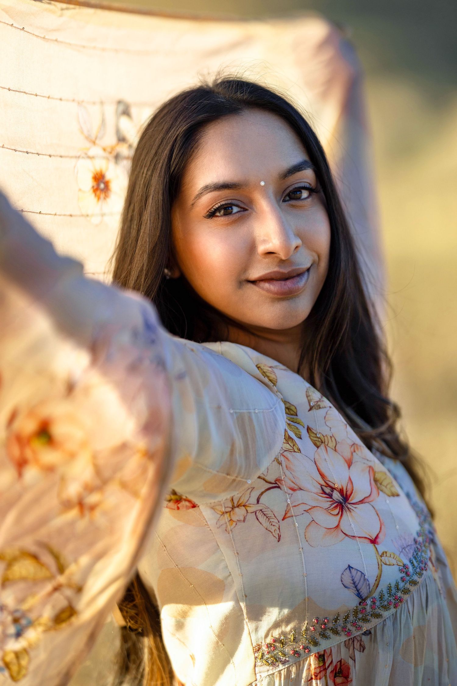 Young Indian woman in floral embroidered dress holds dupatta scarf in golden sunset light, bindi on forehead.