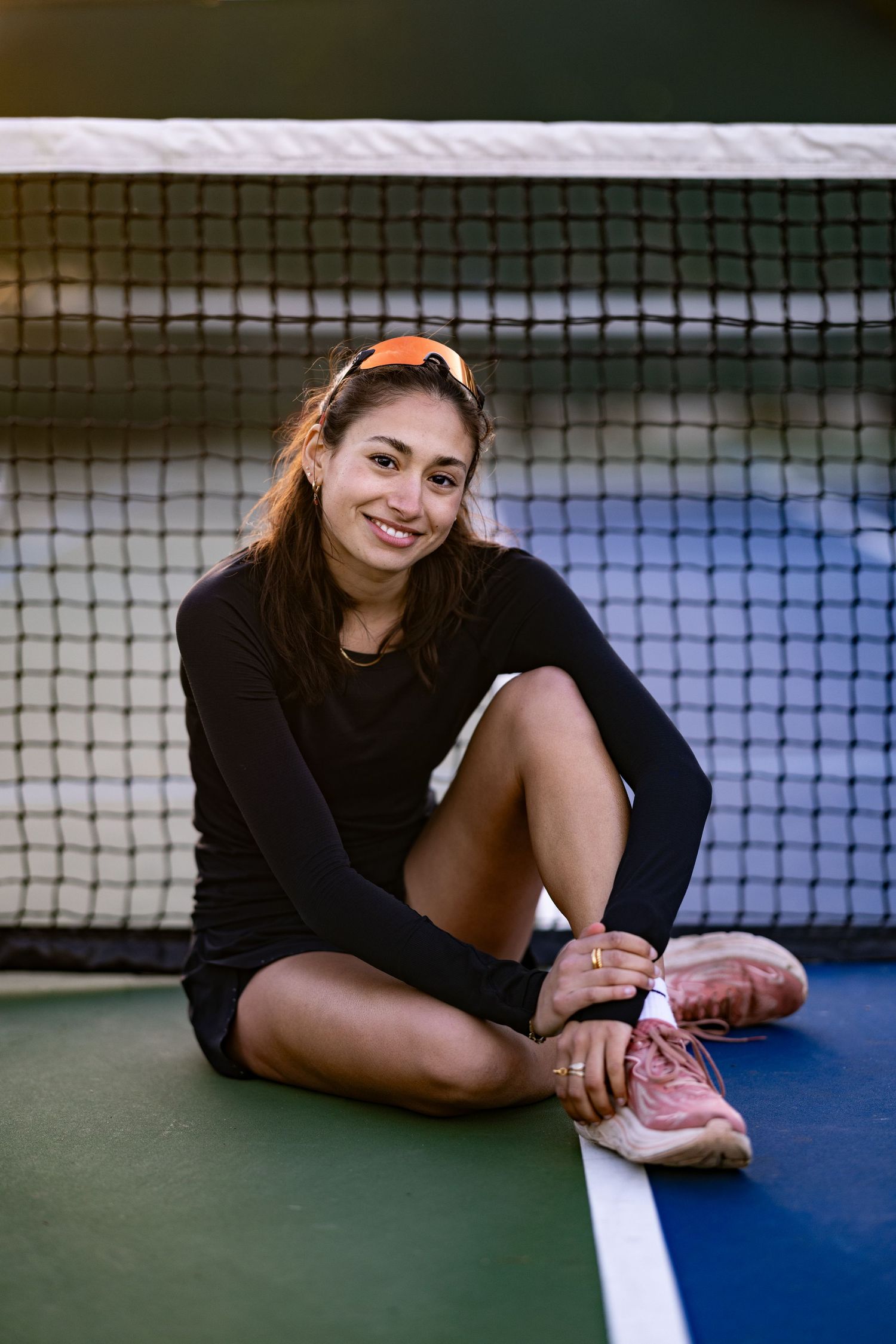 Tennis player in black sweater and pink sneakers sits on court by net during outdoor practice session.
