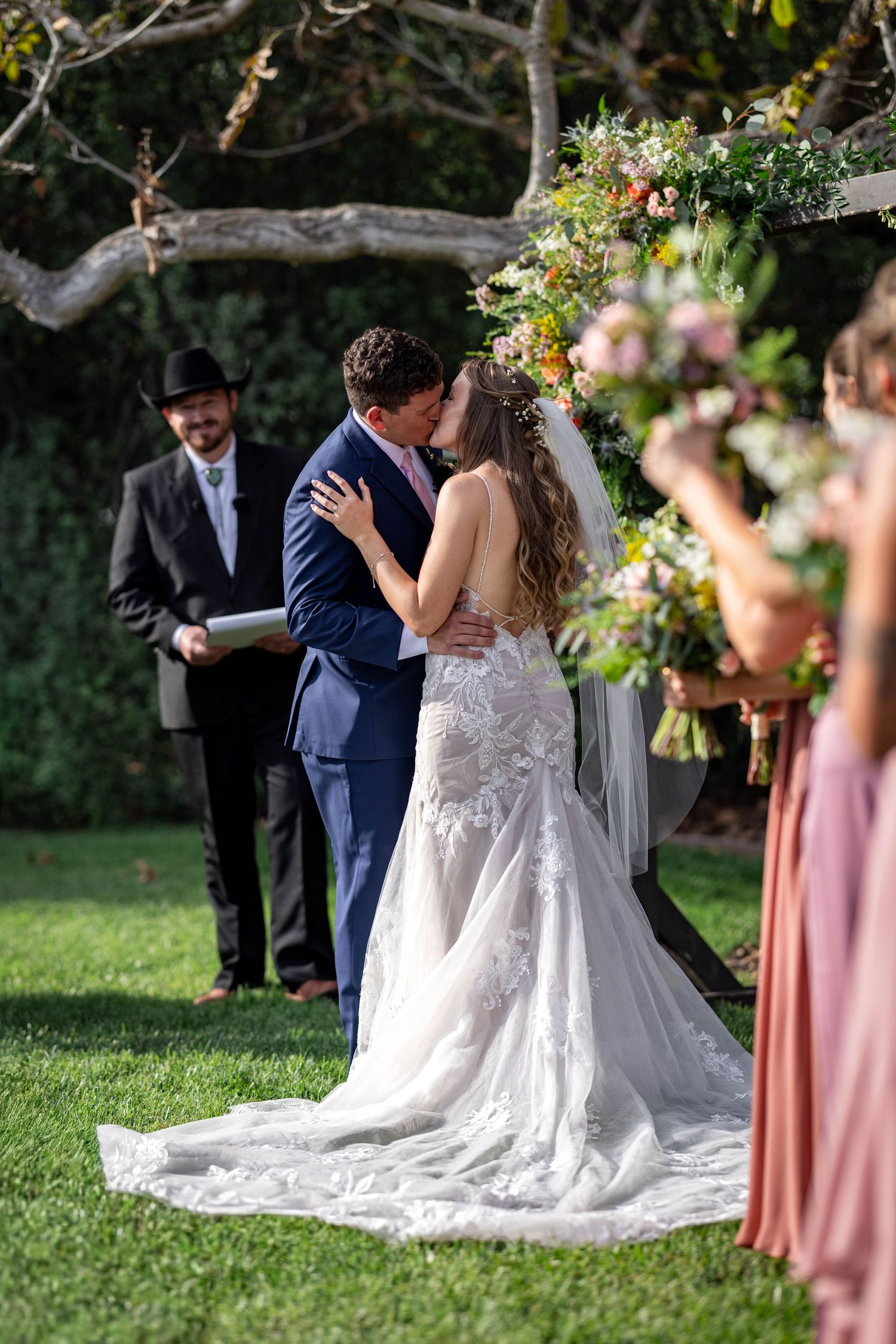 Bride and groom share first kiss at outdoor wedding ceremony with floral arch and officiant in cowboy hat.