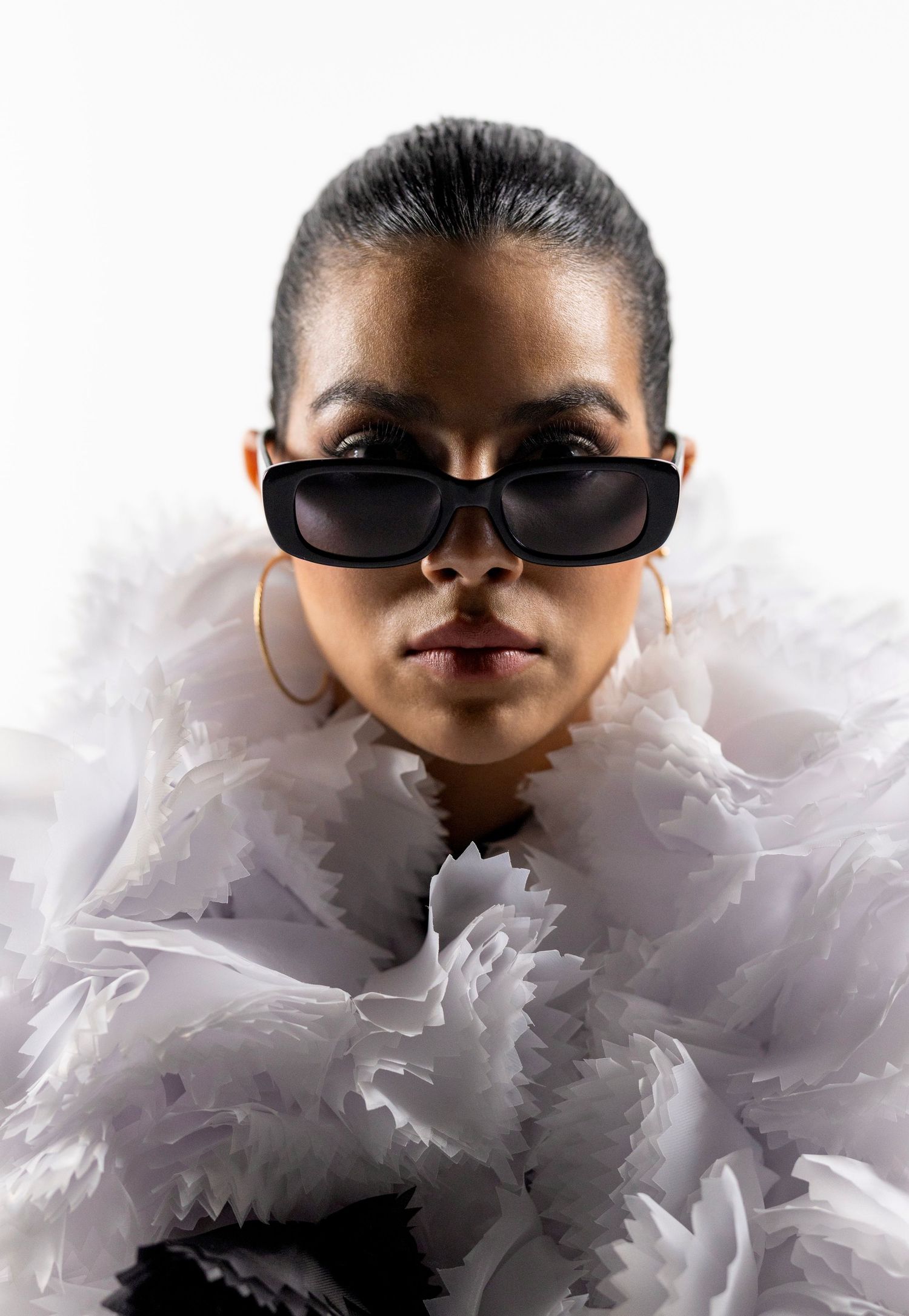 Woman in white ruffled outfit and black rectangular sunglasses with gold hoop earrings poses against white background.
