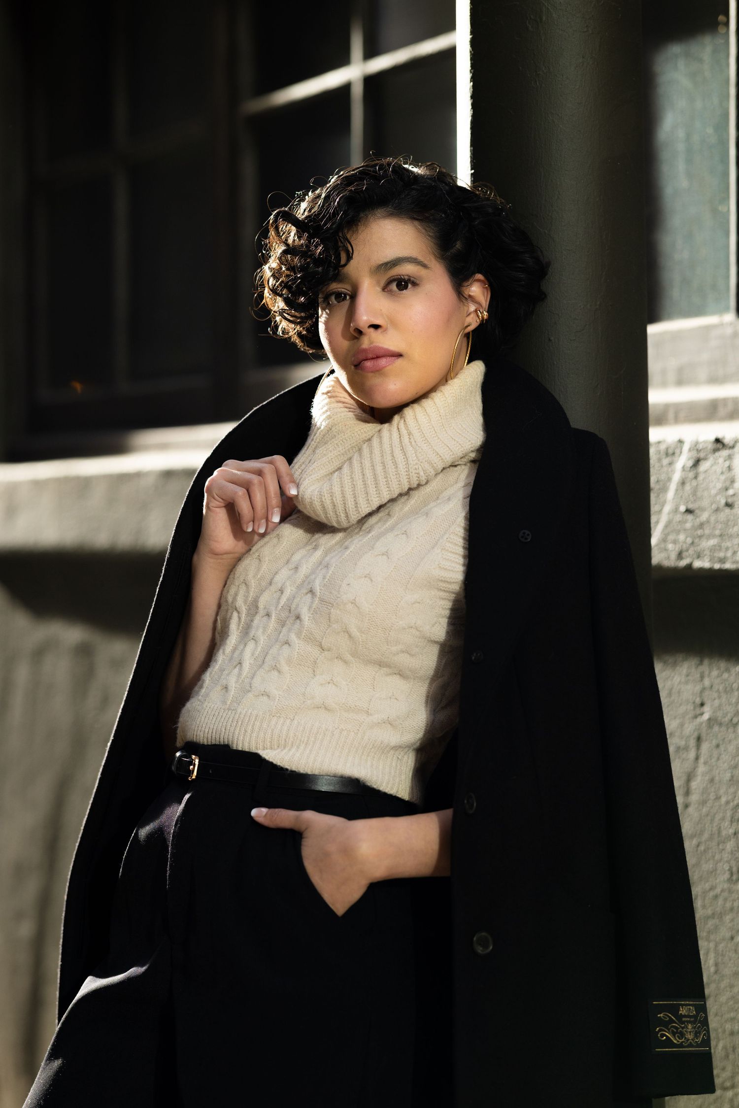 A person in a black coat and cream turtleneck sweater poses against a light wall in a moody portrait series.