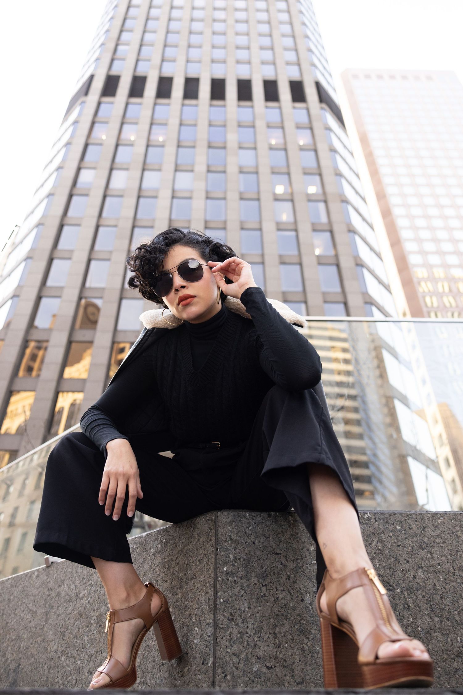 Fashion model in black outfit and sunglasses poses on stone ledge against urban skyscraper backdrop.