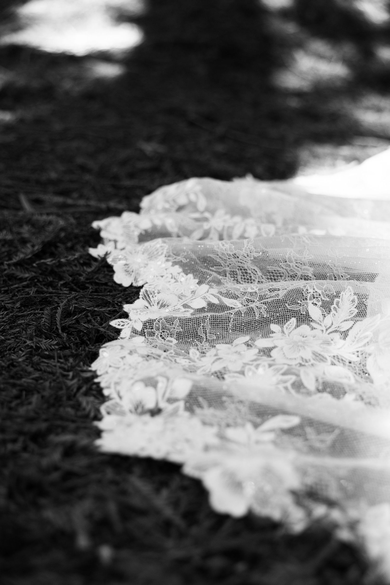 Black and white photo of delicate lace wedding dress train with floral embroidery spread on grass in soft natural light.
