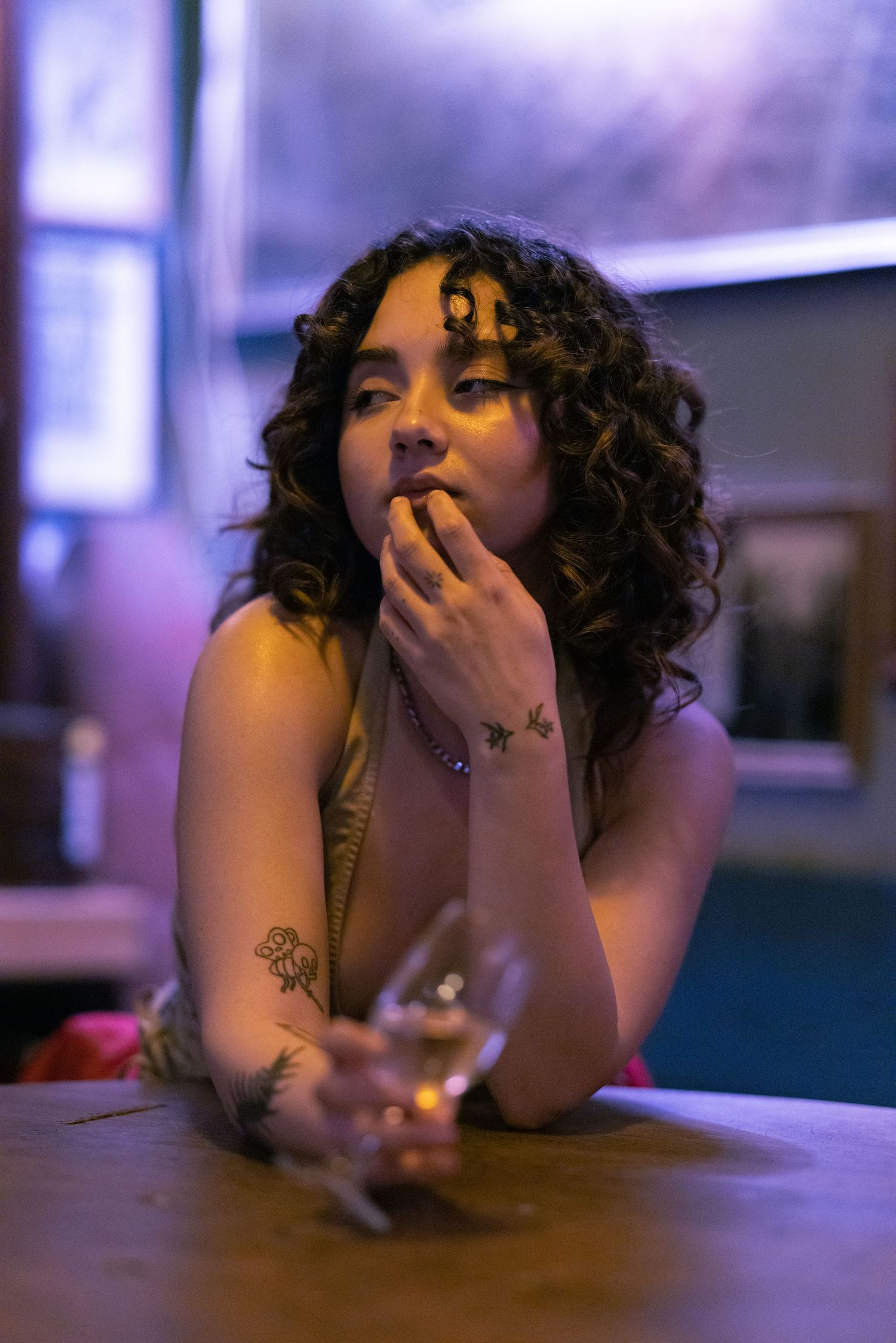 Person with curly hair rests their head on hand at a dimly lit table in moody purple lighting.