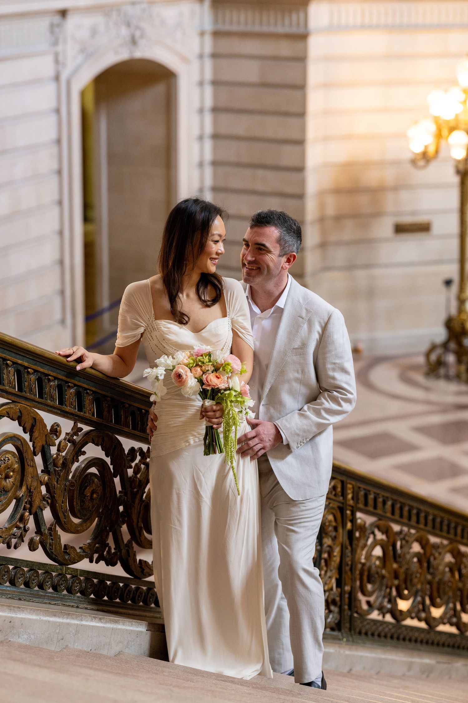 Bride and groom stand on ornate staircase at San Francisco City Hall, she holds pink bouquet, both wearing cream attire.