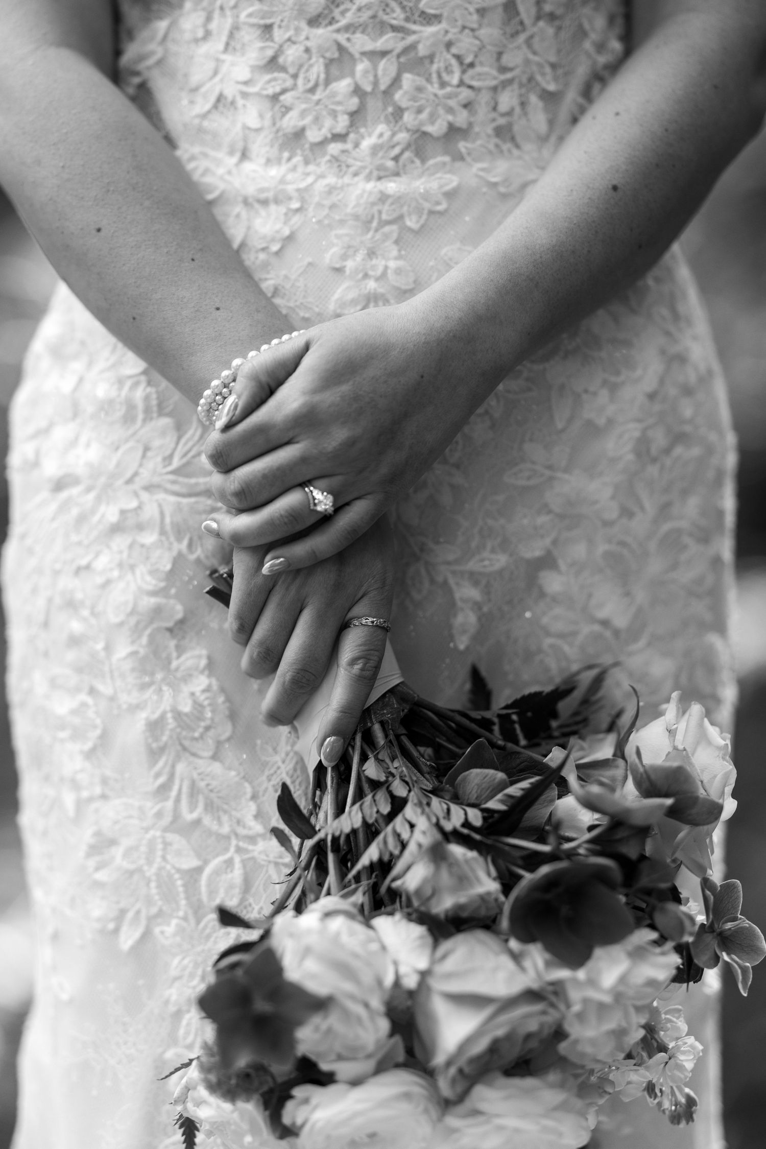 Black and white photo of bride holding bouquet, showing diamond engagement ring and pearl bracelet against lace wedding dress.